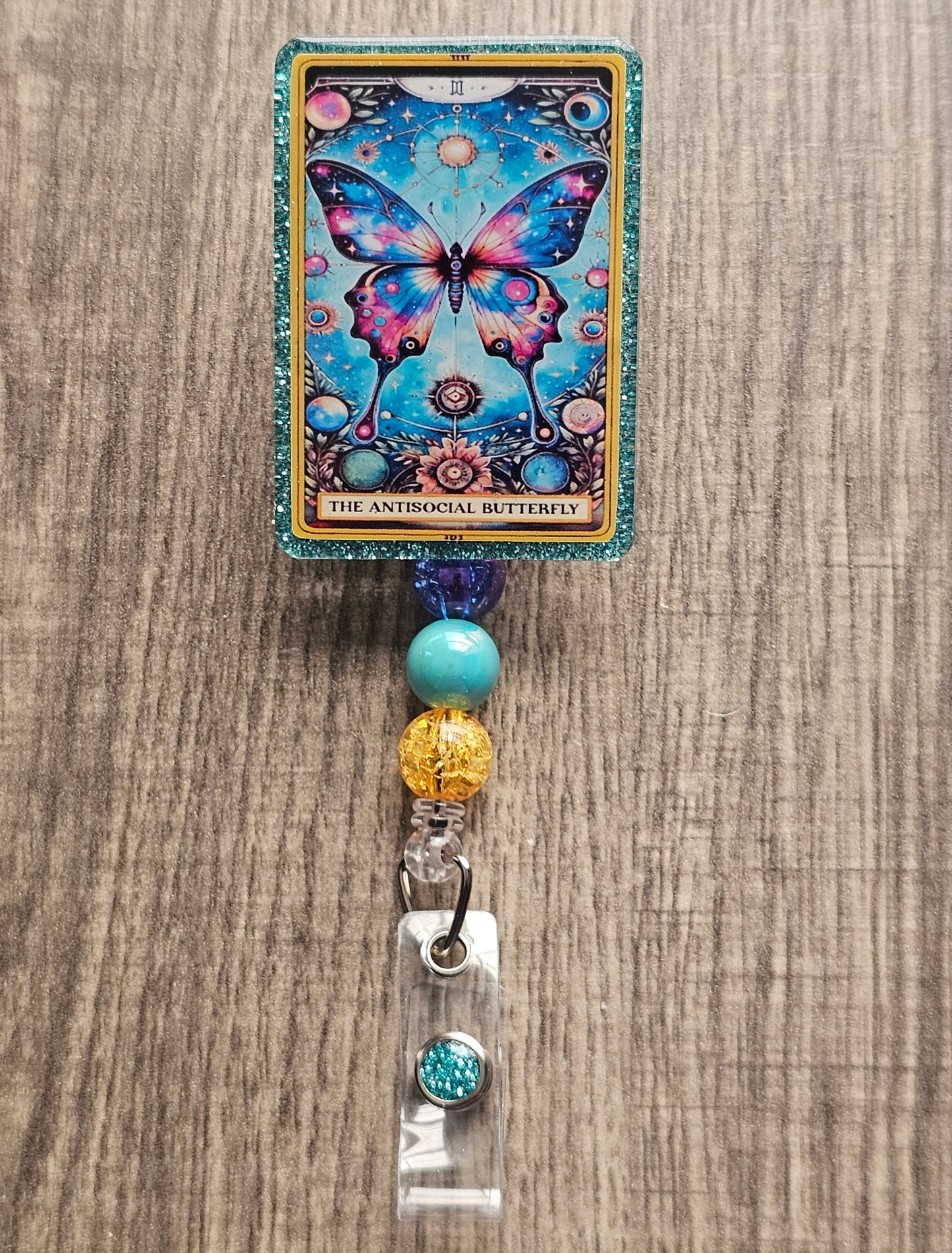 A badge Reel in the shape and design of a tarot card featuring a florescent butterfly surrounded by a magical space background. With the badge holder being titled "The Antisocial Butterfly."