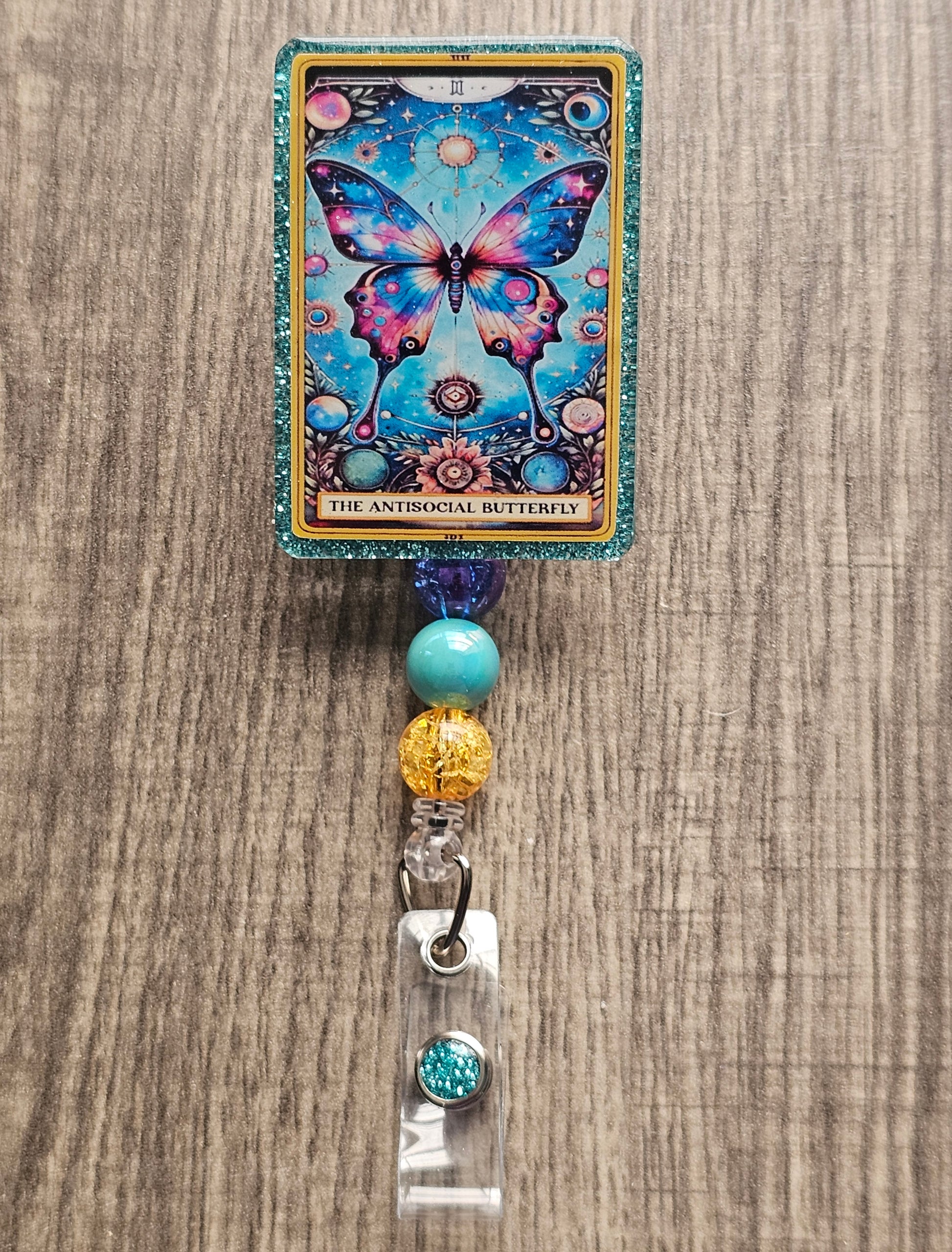 A badge Reel in the shape and design of a tarot card featuring a florescent butterfly surrounded by a magical space background. With the badge holder being titled "The Antisocial Butterfly."