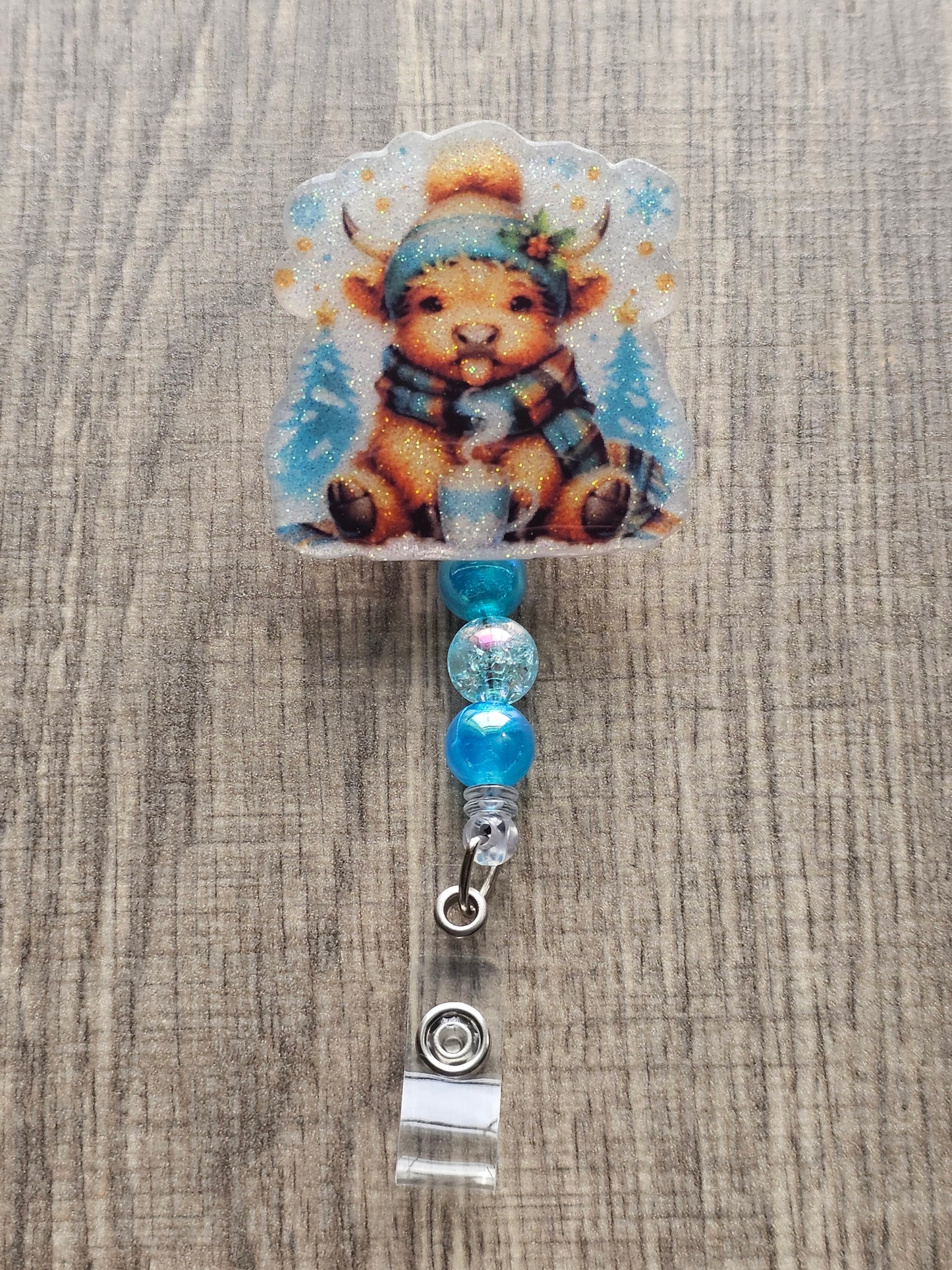 A badge reel featuring an adorable highland cow playfully sticking its tongue out, this design is complete with a warm hat, cozy scarf, and a steaming cup of hot cocoa.