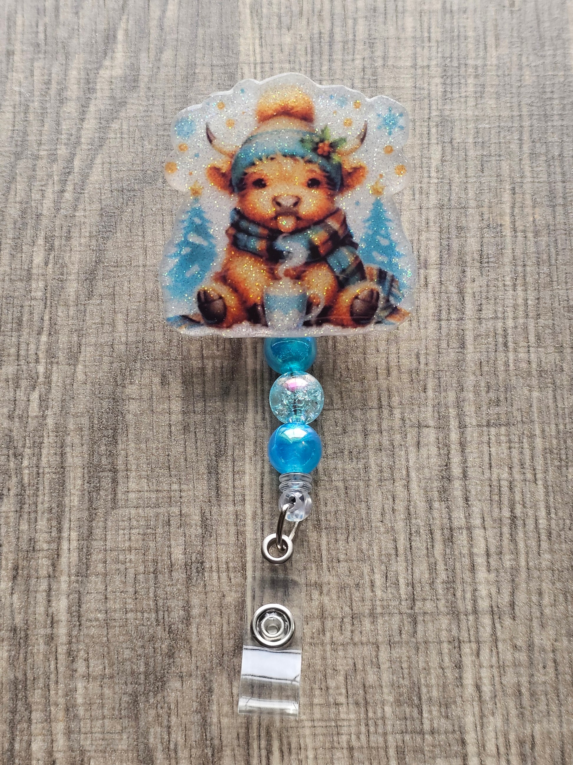 A badge reel featuring an adorable highland cow playfully sticking its tongue out, this design is complete with a warm hat, cozy scarf, and a steaming cup of hot cocoa.