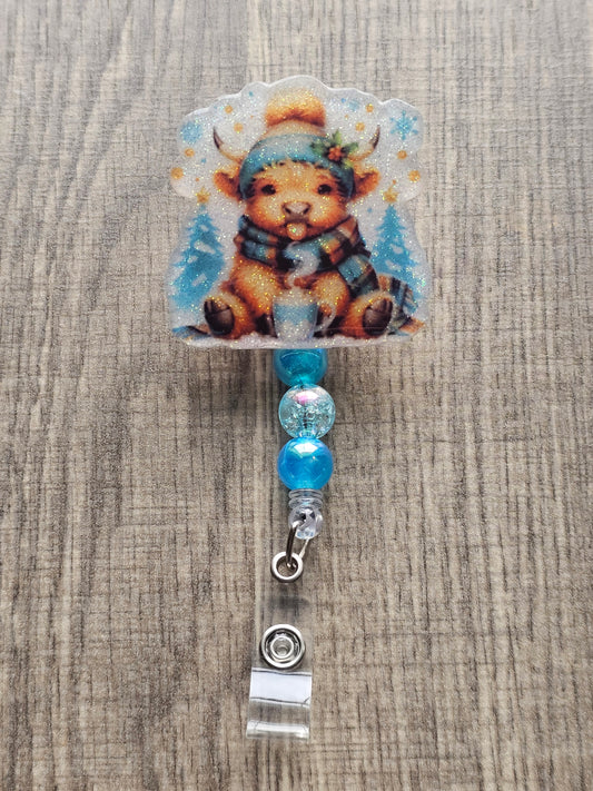A badge reel featuring an adorable highland cow playfully sticking its tongue out, this design is complete with a warm hat, cozy scarf, and a steaming cup of hot cocoa.