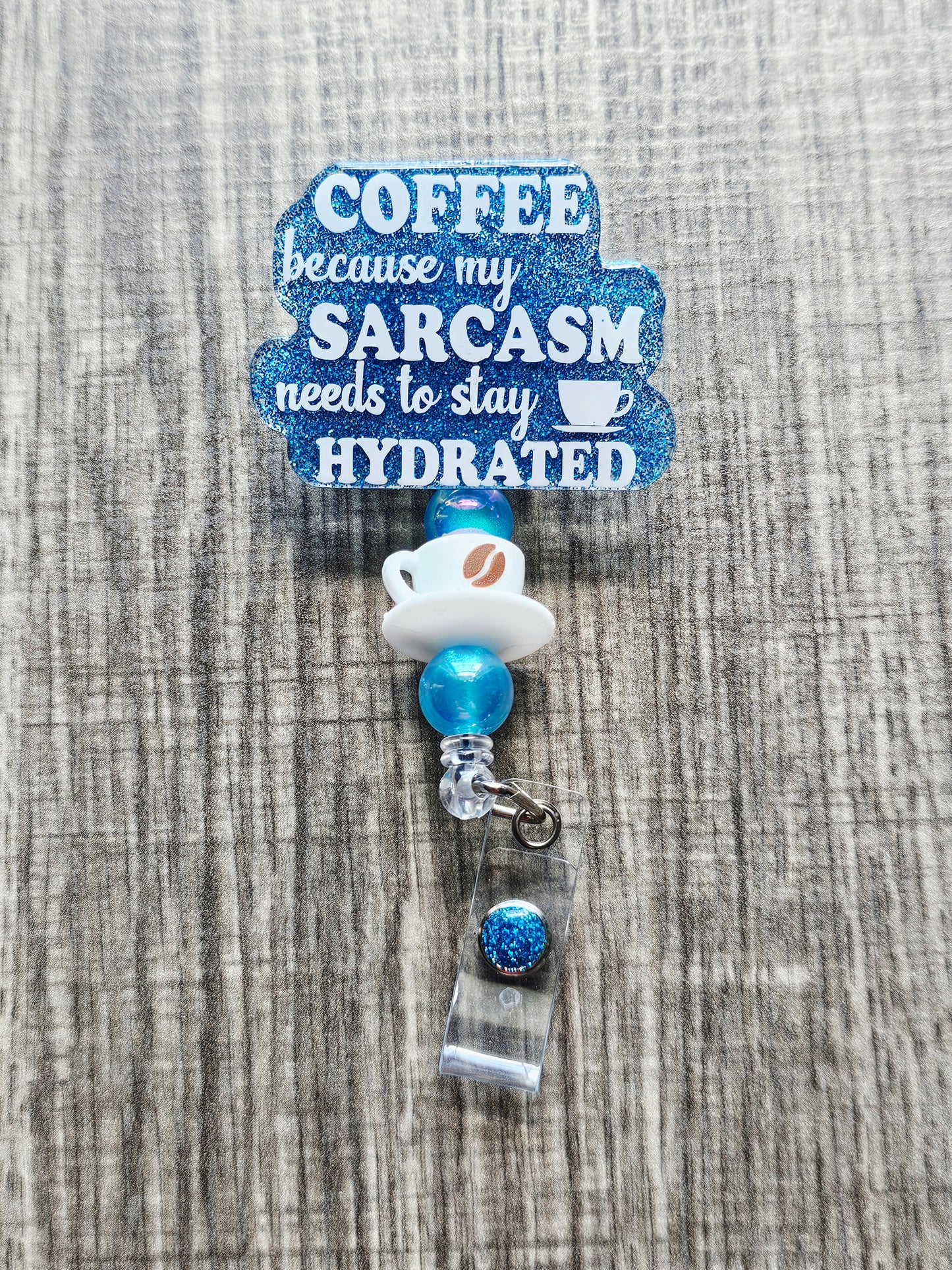 Funny coffee badge reel stating "Coffee because my sarcasm needs to stay hydrated" with a coffee cup bead.