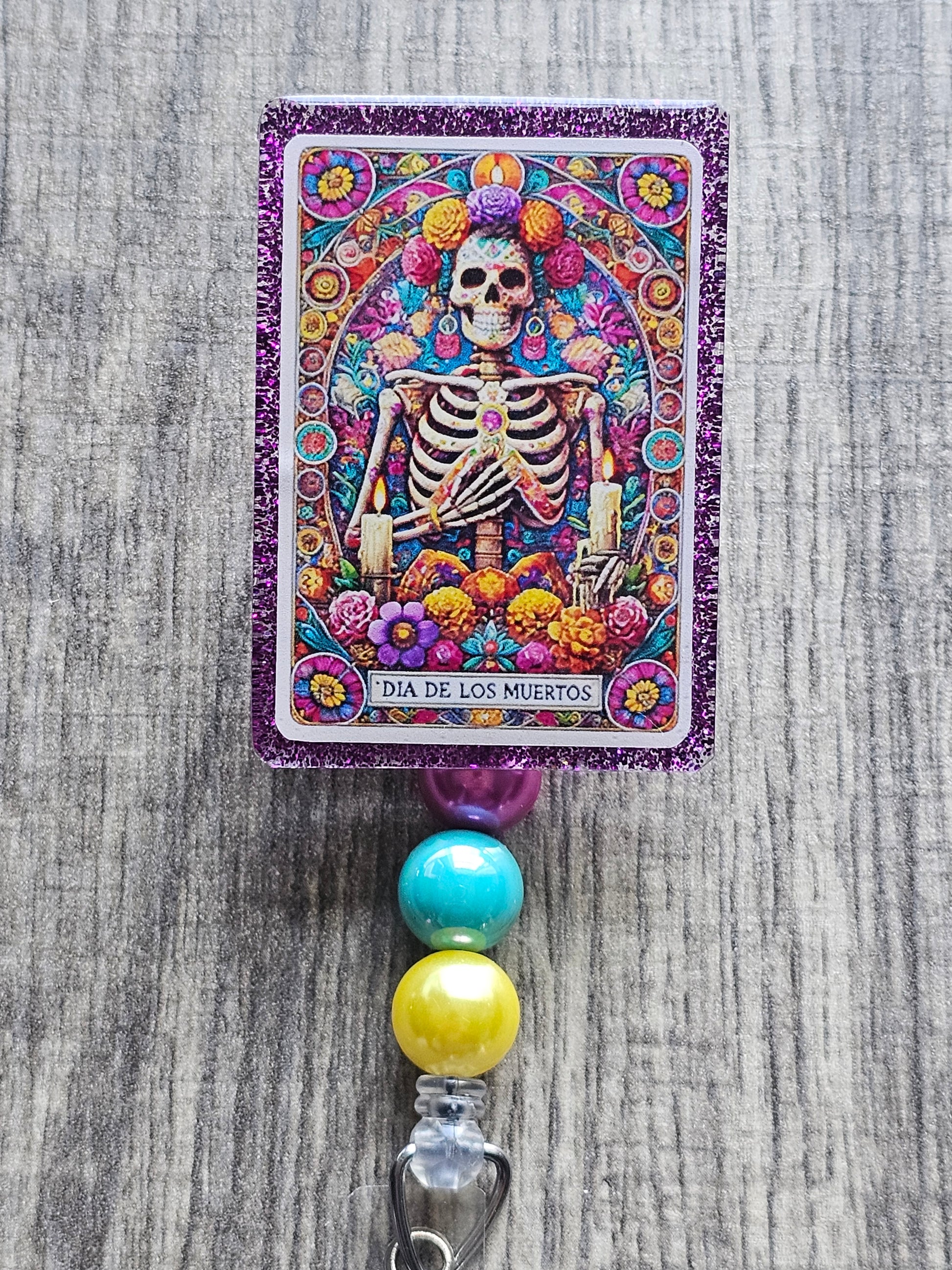 a badge reel that displays a skeleton holding their hand towards their chest while being surrounded by a wide variety of flowers along with a flower crown in the design of a tarot card