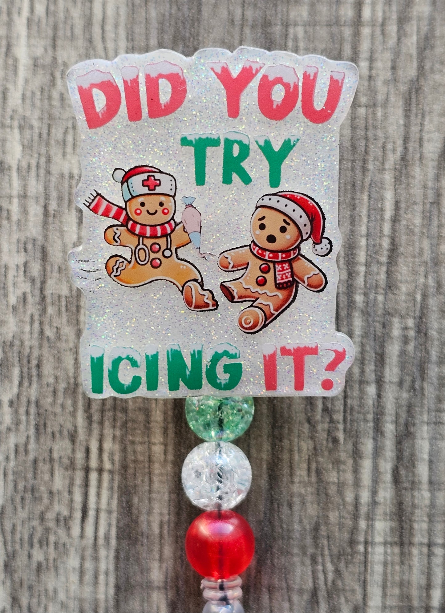 A Badge reel stating Did You Try Icing It? while displaying a gingerbread man adorned with a nurse Christmas hat and stethoscope, while the other gingerbread man sits on the floor sad adorned with a Christmas hat and scarf along with a broken foot.