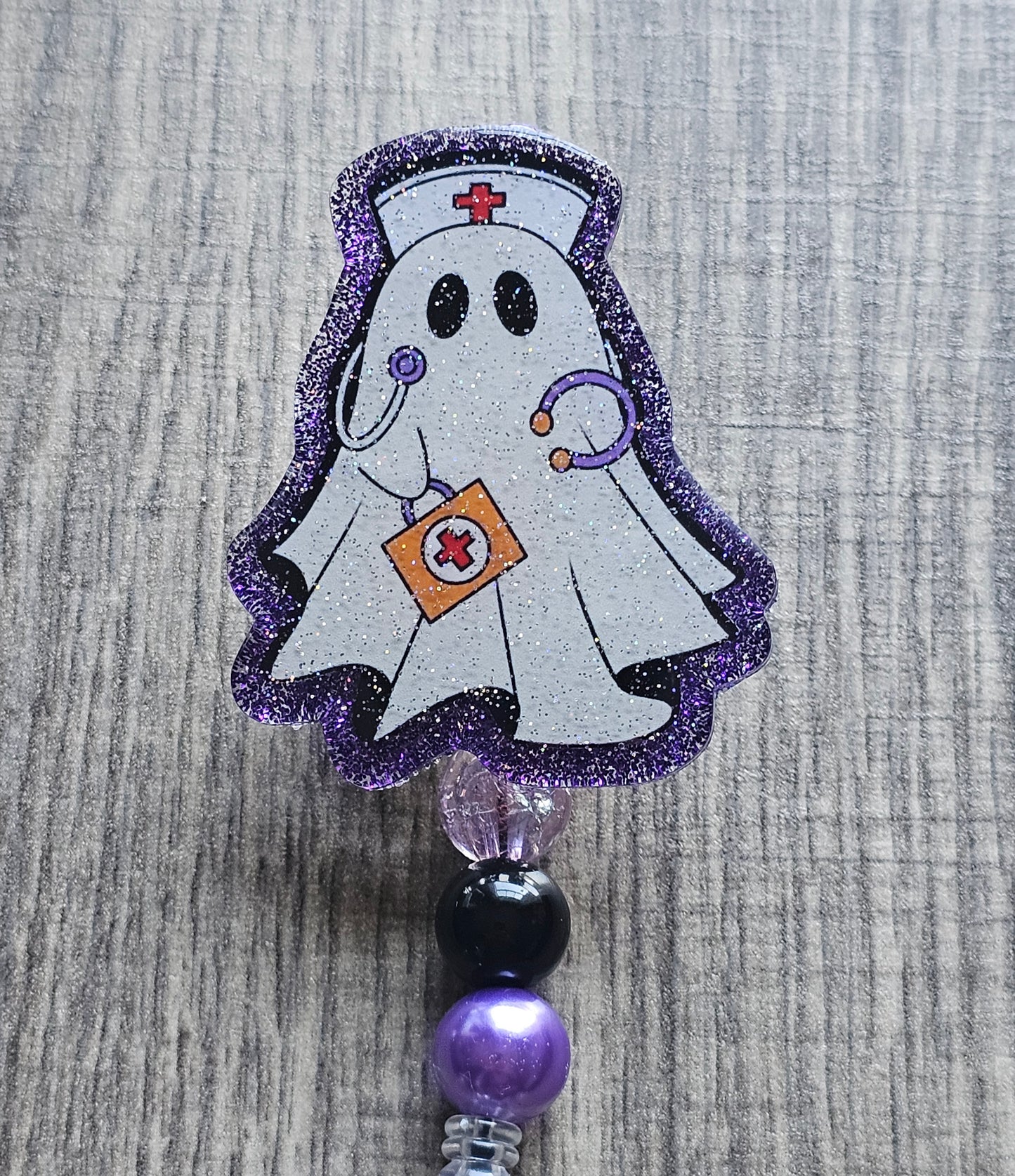 a badge reel shaped as a ghost with the design of a ghost nurse with a nursing hat, stethoscope along with a first aid kit