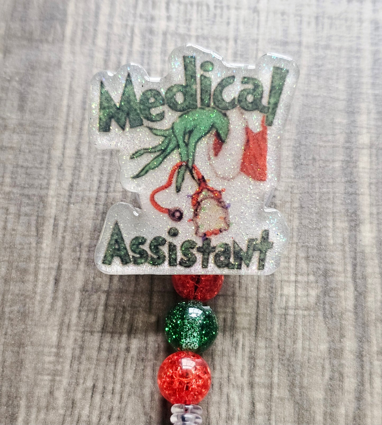 Badge reel depicting the Grinch's hand in a festive sweater holding stethoscopes, with the words "Medical Assistant" in a Christmas theme.