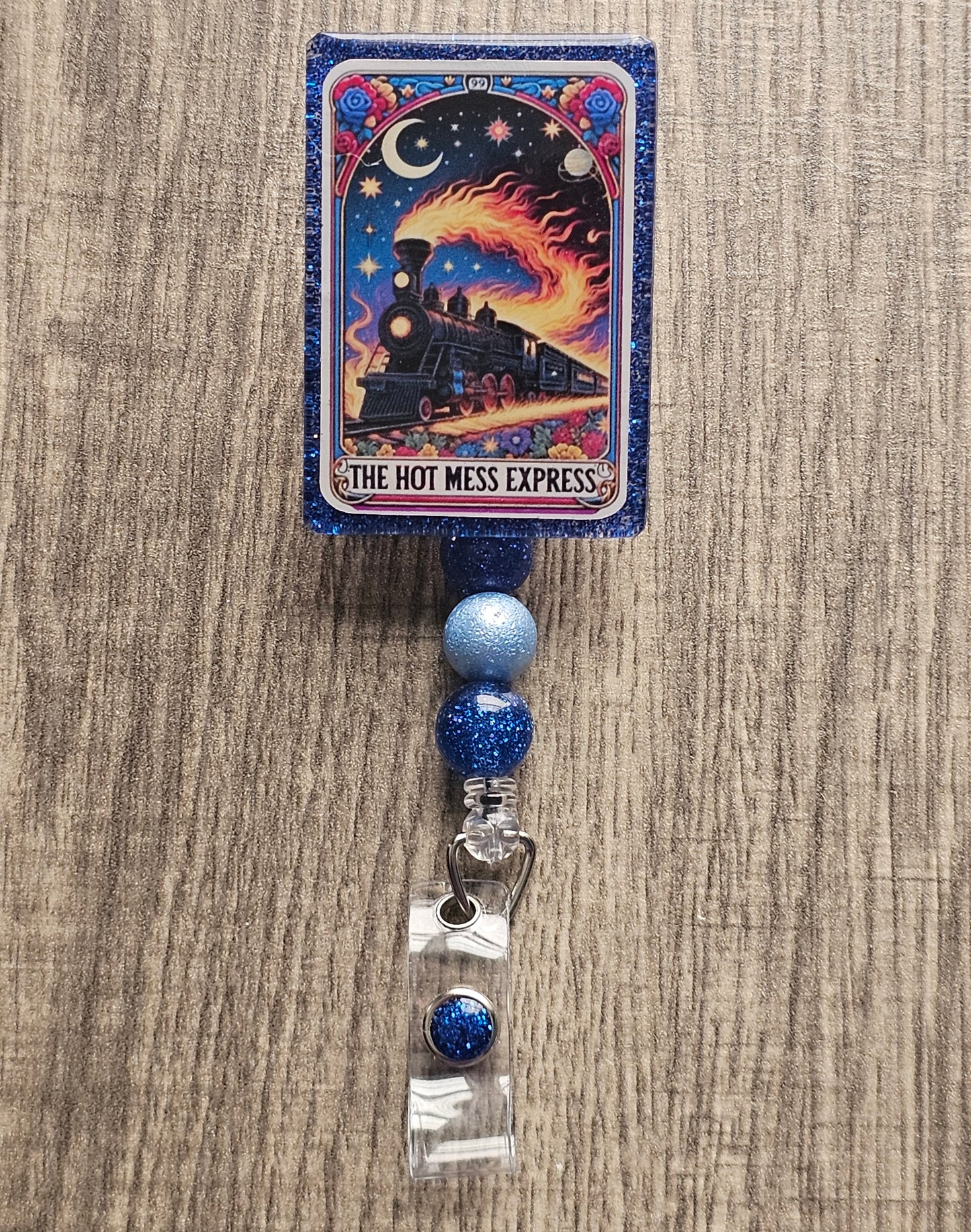 A badge reel in the shape of a tarot card featuring a flaming train going across some tracks with space in the background, with the badge holder being titled "The Hot Mess Express."