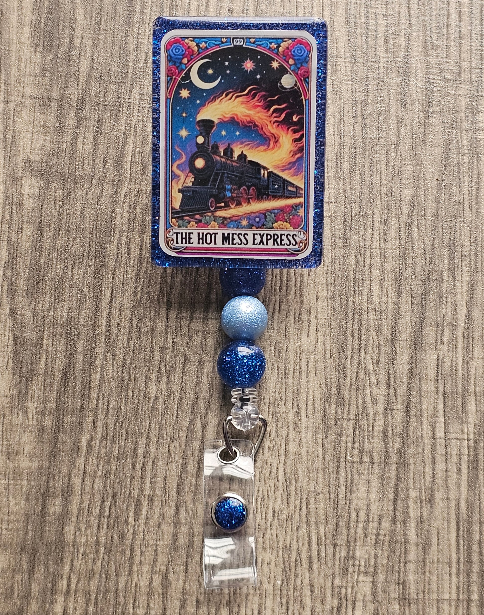 A badge reel in the shape of a tarot card featuring a flaming train going across some tracks with space in the background, with the badge holder being titled "The Hot Mess Express."