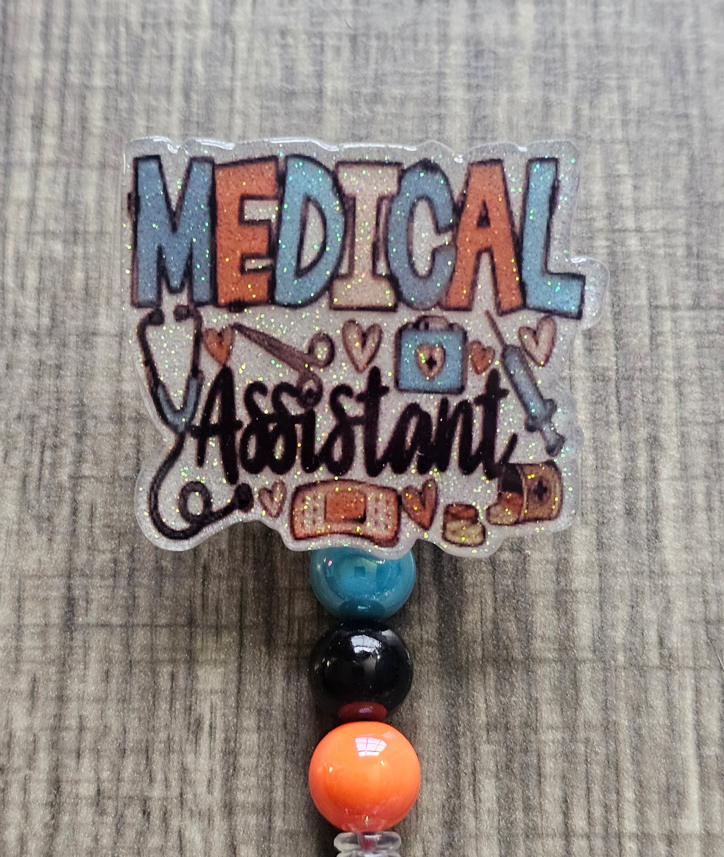 White glitter acrylic badge reel with "Medical Assistant" text, featuring a stethoscope, scissors, first aid kit, syringe, hearts, and medicine, with creatively matching decorative beads.