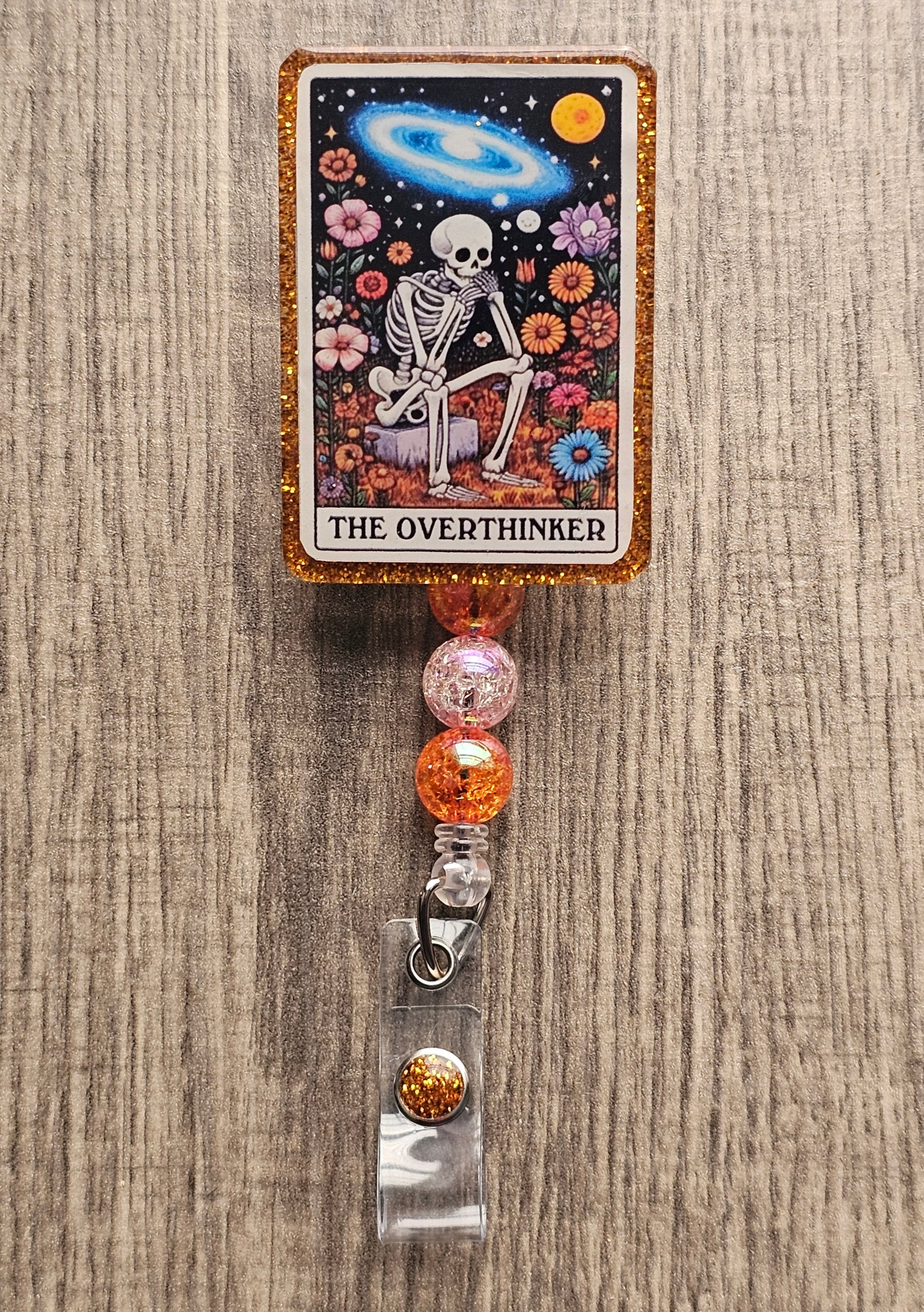 Badge Reel Shaped as a tarot card featuring a skeleton sitting on a rock surrounded by flowers and space thinking with the badge reel being titled the Overthinker