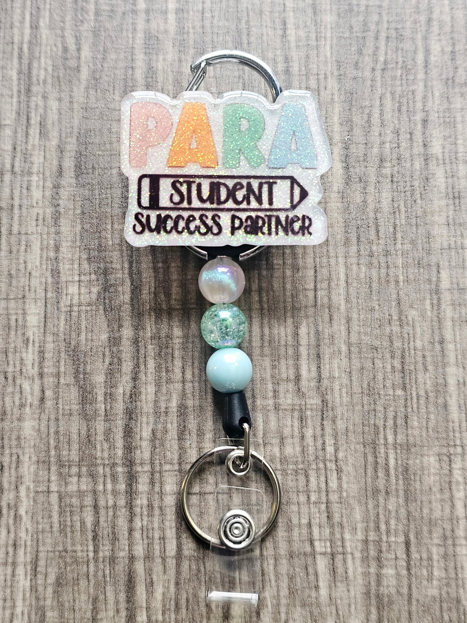 White glitter acrylic badge reel with "PARA," "Student" within a pencil, and "Success Partner" below, with well-coordinating decorative beads.