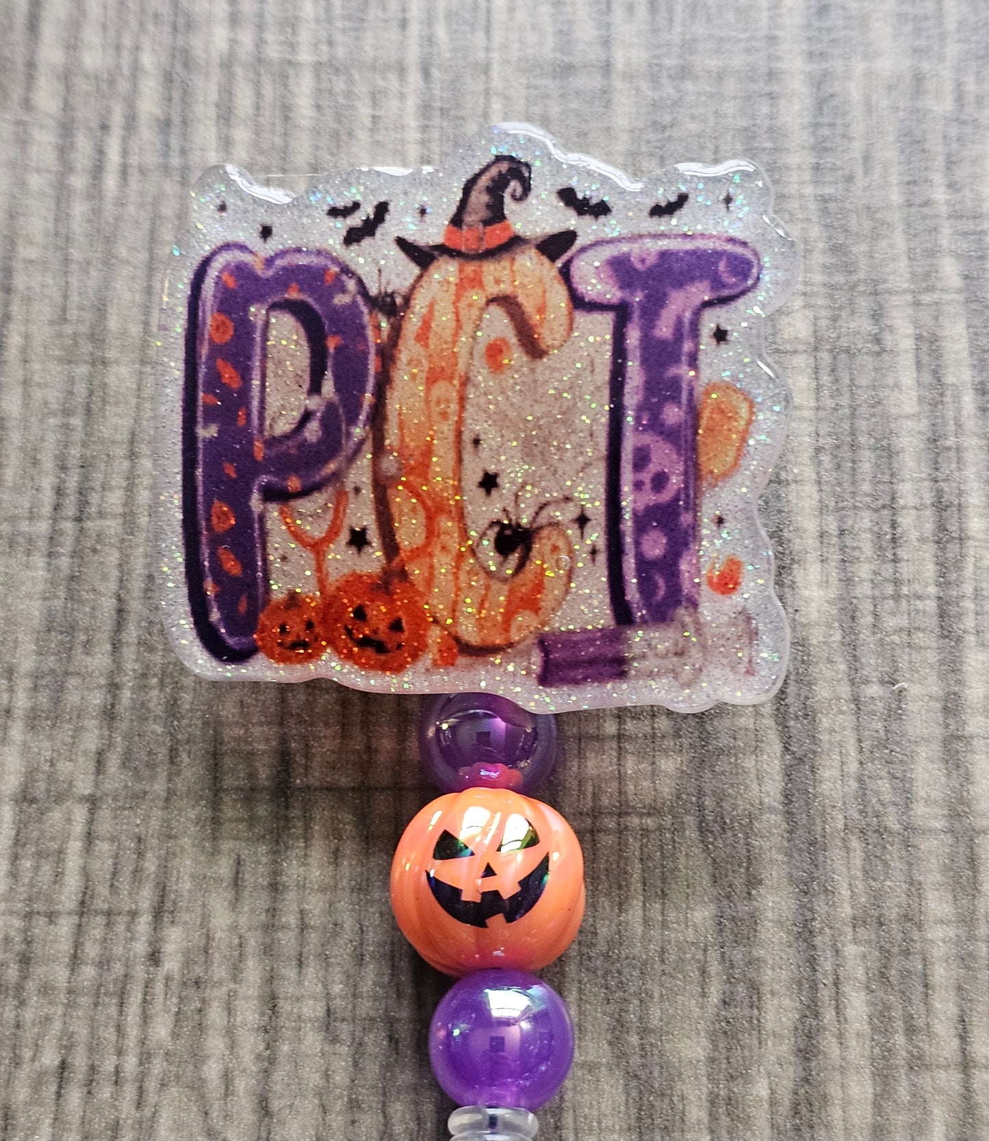 White glitter acrylic badge reel with Halloween-themed "PCT" letters, featuring bats, jack-o-lanterns, a Halloween syringe, and a witch's hat on the 'C', with matching beads and a jack-o-lantern bead.