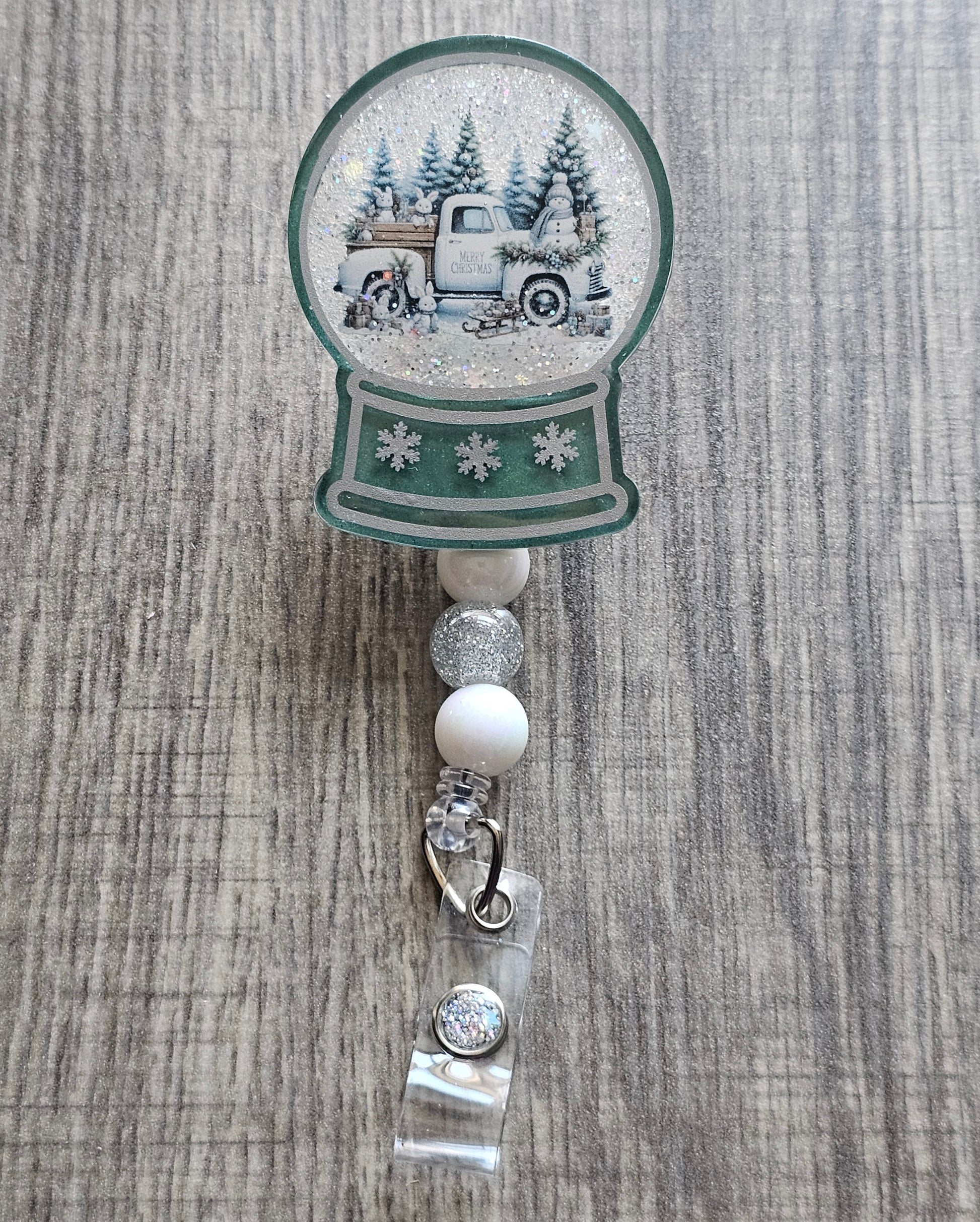 A badge reel with a snow globe design featuring a snowed in tuck surrounded by Christmas Tress, and snowmen with the words Merry Christmas on the side of the door.