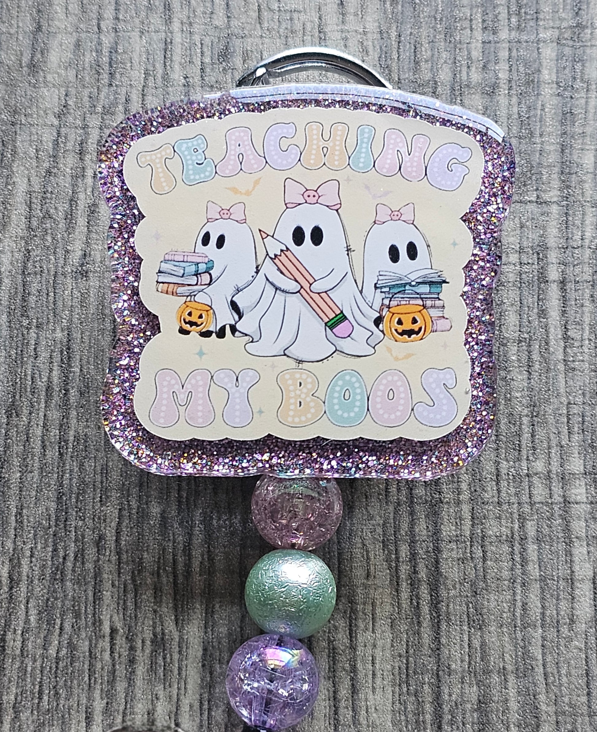 a badge reel stating teaching my boos with the ghosts wearing bows incorporated along the design; one holding a pencil while the others have books along with a trick or treat jar