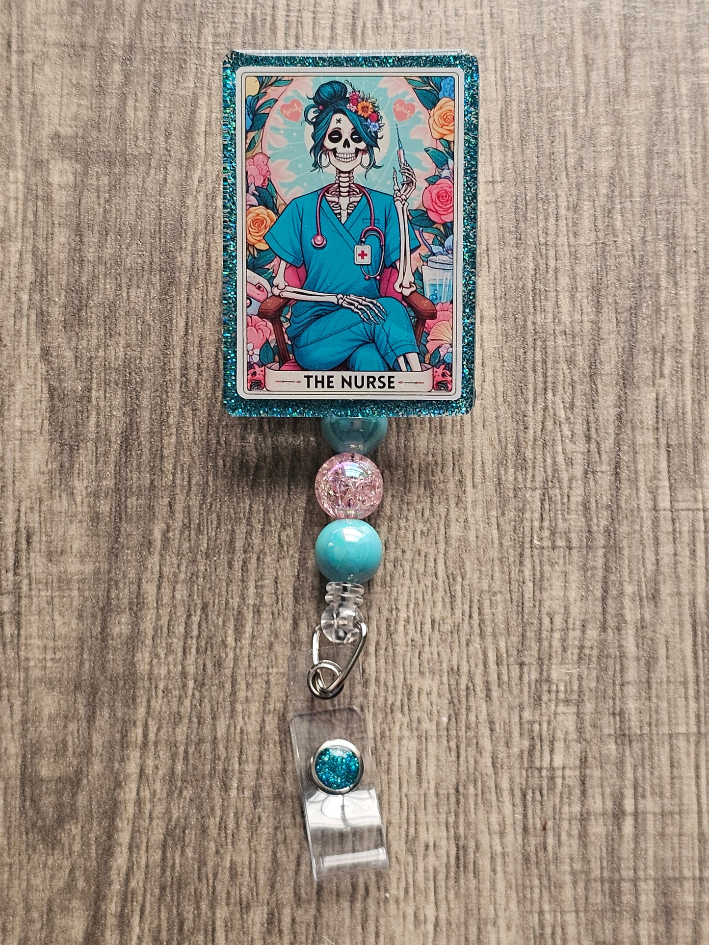 A badge Reel in the shape and design of a tarot card featuring a a skeleton nurse in scrubs sitting with her legs crossed with a syringe in her hand along with a stethoscope around her neck surrounded by flowers. With the badge holder title being "The Nurse."