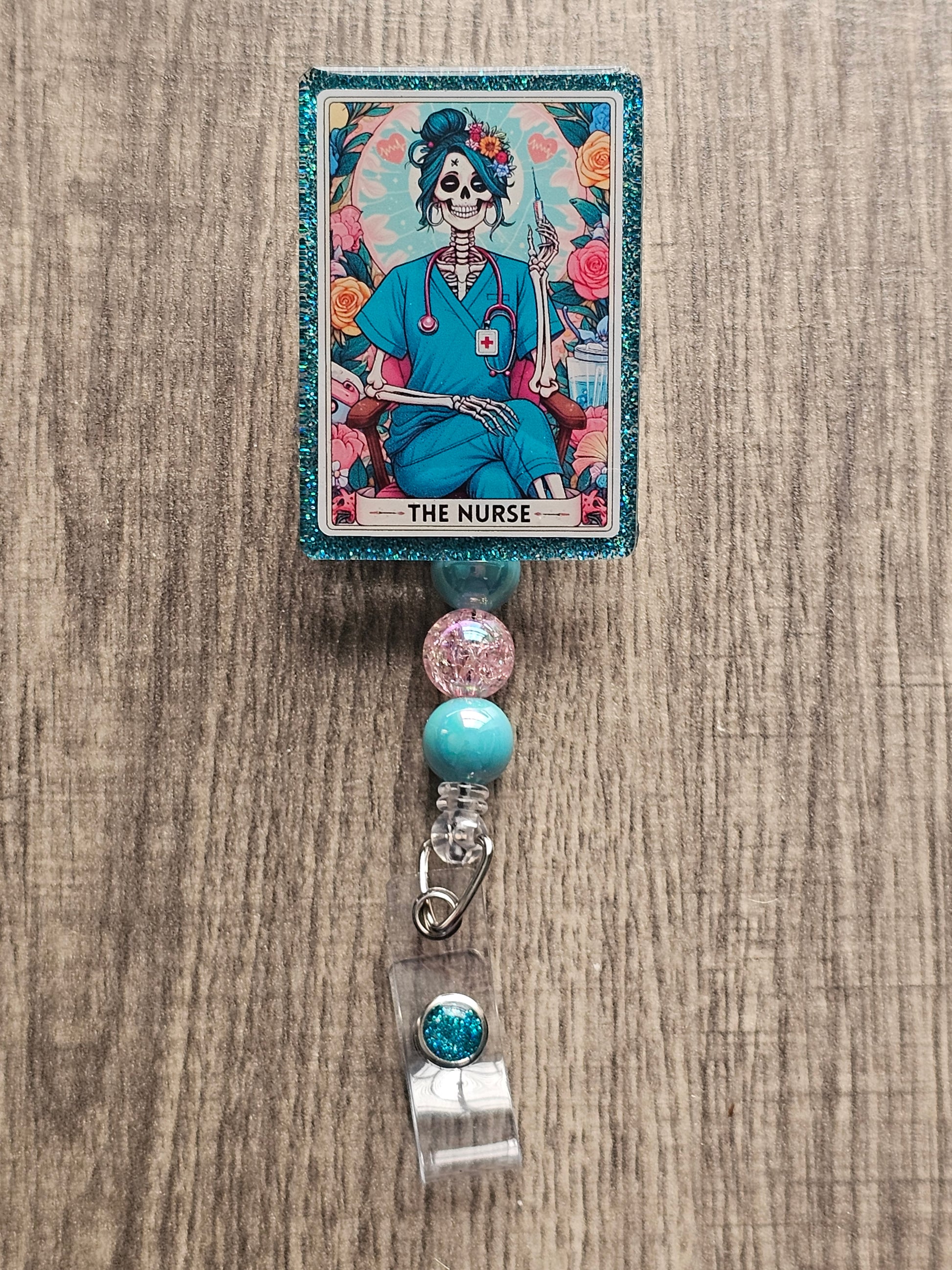 A badge Reel in the shape and design of a tarot card featuring a a skeleton nurse in scrubs sitting with her legs crossed with a syringe in her hand along with a stethoscope around her neck surrounded by flowers. With the badge holder title being "The Nurse."