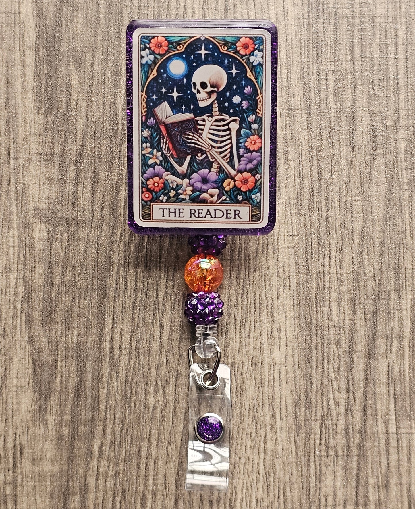 Features a badge reel in the shape of a tarot Card featuring a skeleton reading a book while being surrounded by flowers with the title of the badge holder being "The Reader"
