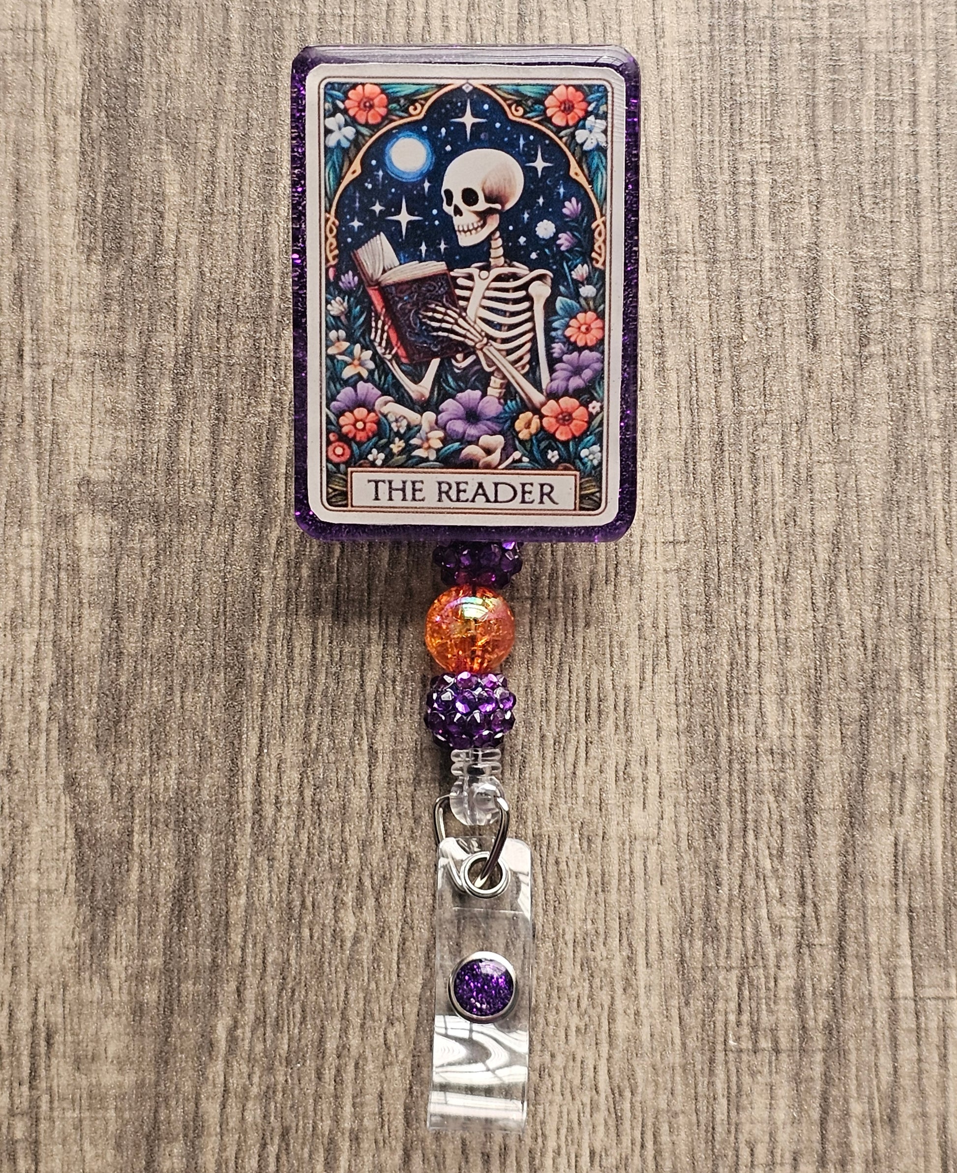 Features a badge reel in the shape of a tarot Card featuring a skeleton reading a book while being surrounded by flowers with the title of the badge holder being "The Reader"