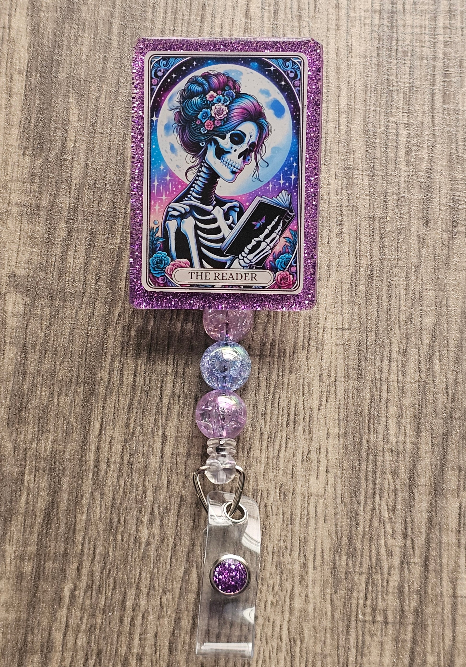 A Badge Reel Shaped as a tarot Card featuring a skeleton of a woman reading a book, with the moon in the background. The Badge Holder being title The reader