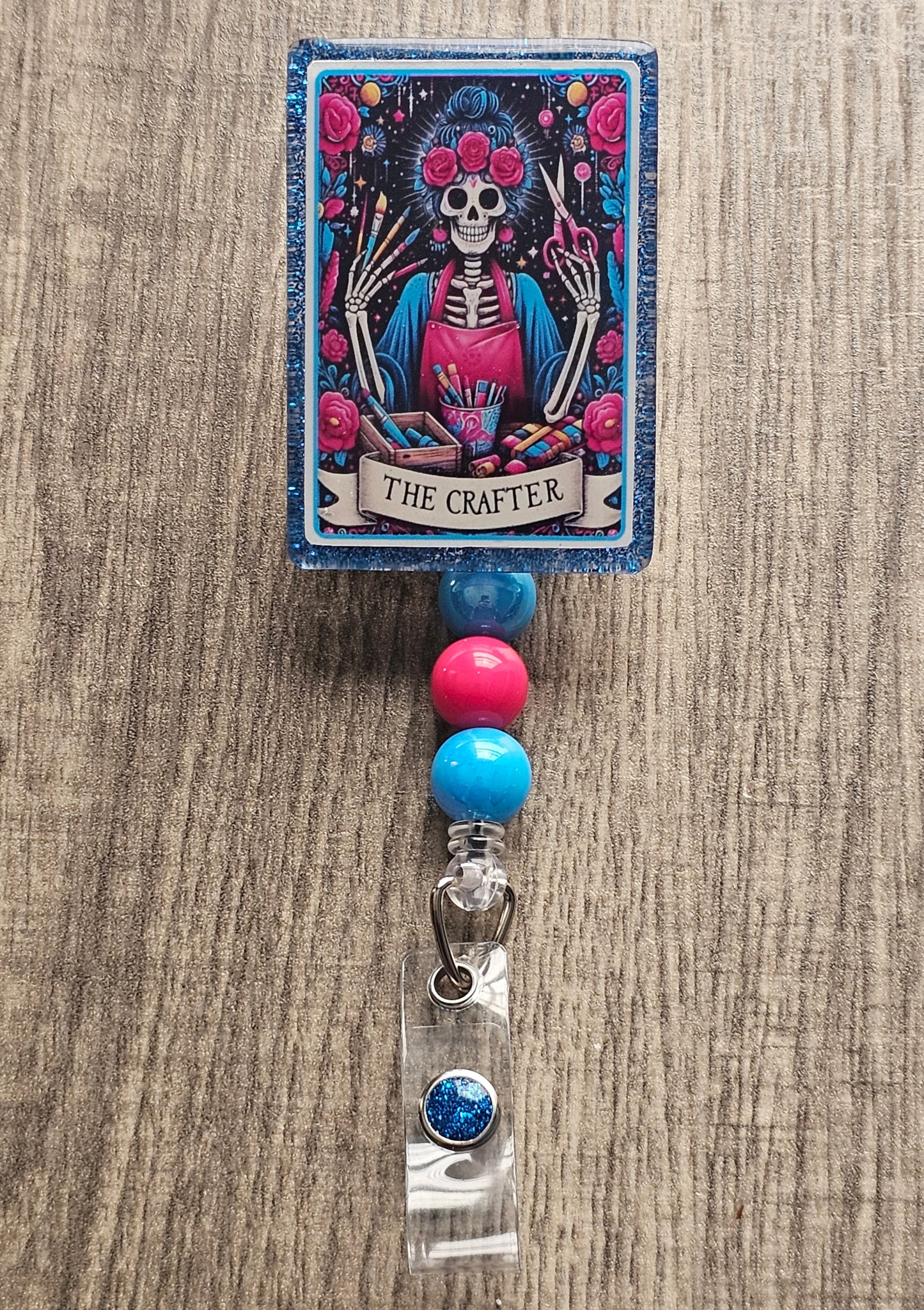 A badge Reel in the shape and design of a tarot card featuring a crafty skeleton woman with an apron surrounded by flowers while holding scissors and painting accessories in her hands along with a floral crown. The Badge Holder being titled The Crafter.