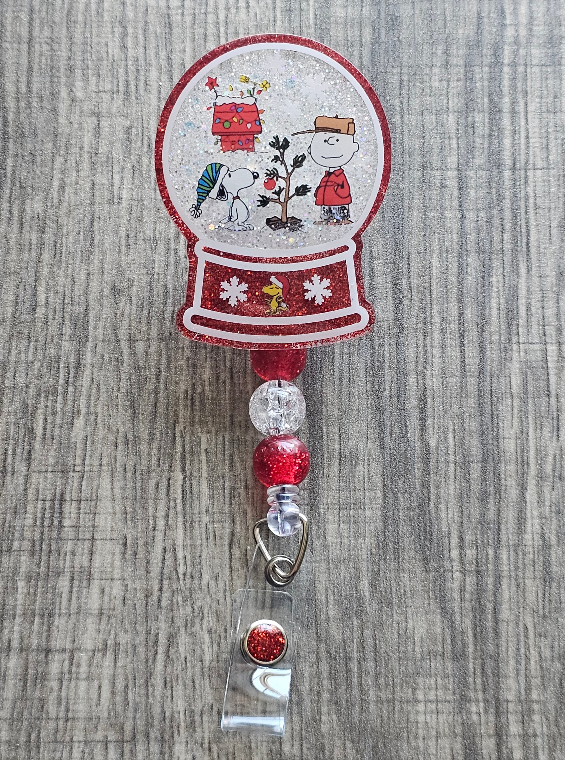A badge reel with a snow globe design featuring peanuts and snoopy next to a tree with a festive house behind them surrounded with snow.