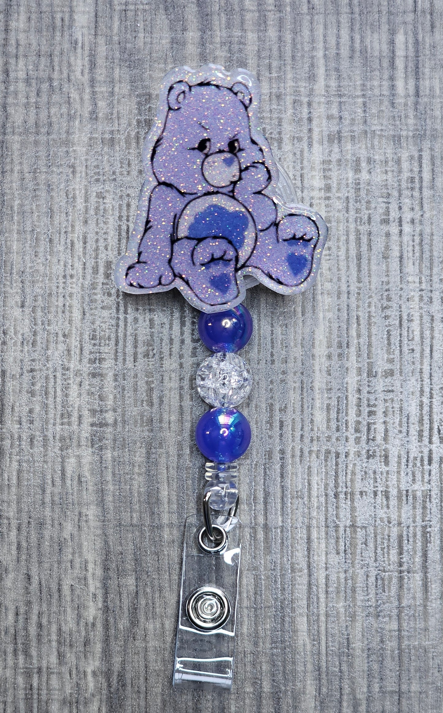 Grumpy Care Bear-shaped badge holder with blue and silver beads on a textured gray background