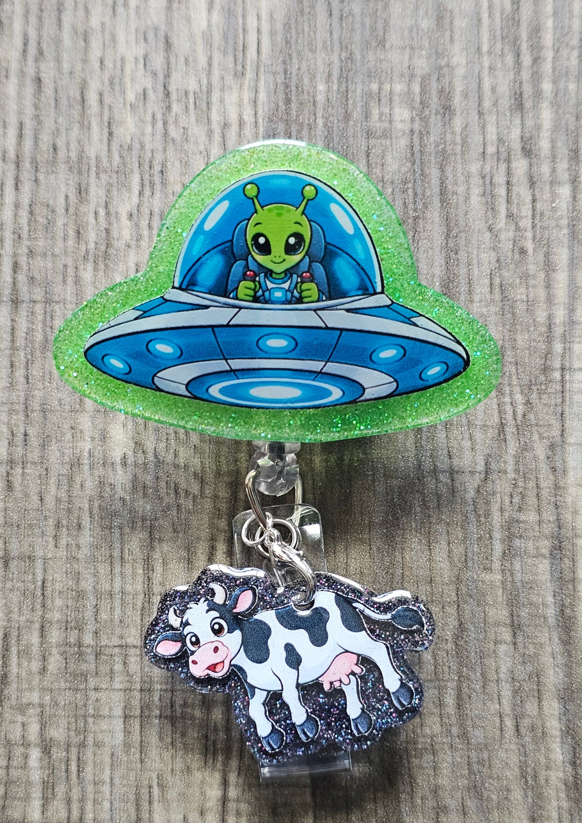 Quirky retractable alien badge reel showing a silver UFO spaceship abducting a small cow figurine attached to the lower ID clip, illustrating a classic sci-fi tractor beam scene.