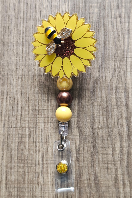 A badge reel with a sunflower design, featuring a yellow flower with brown center, clear body, a bee, and decorative elements like beads.
