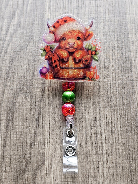 A badge reel that showcases an adorable highland cow nestled in a bucket, wrapped in a cozy blanket, adorned with vibrant flowers, Christmas ornaments, and charmingly wrapped gifts. Topped with a plaid Christmas hat.