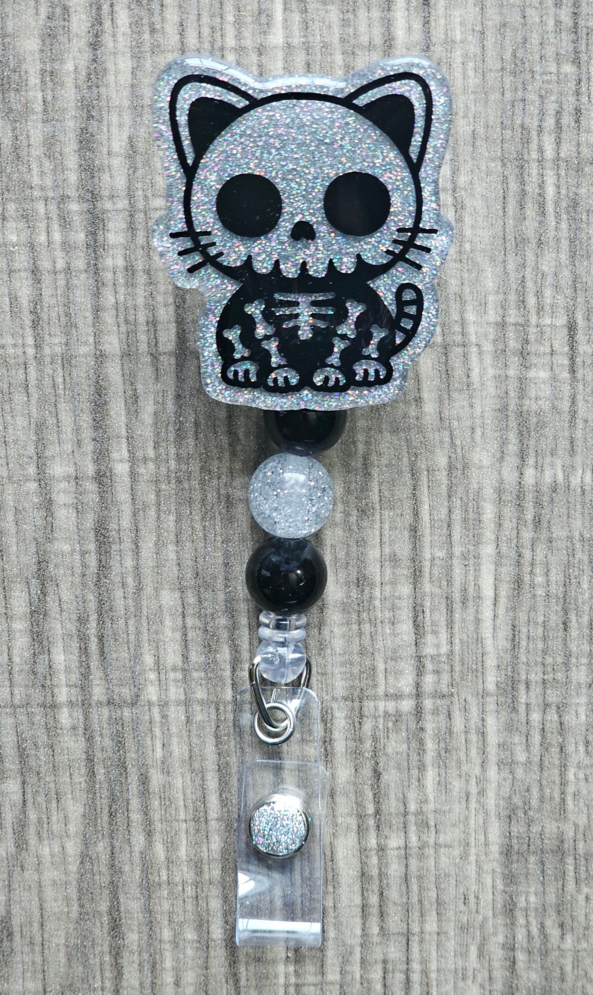 Retractable badge reel featuring a black and silver glittery skeleton cat acrylic topper with decorative black and silver beads.