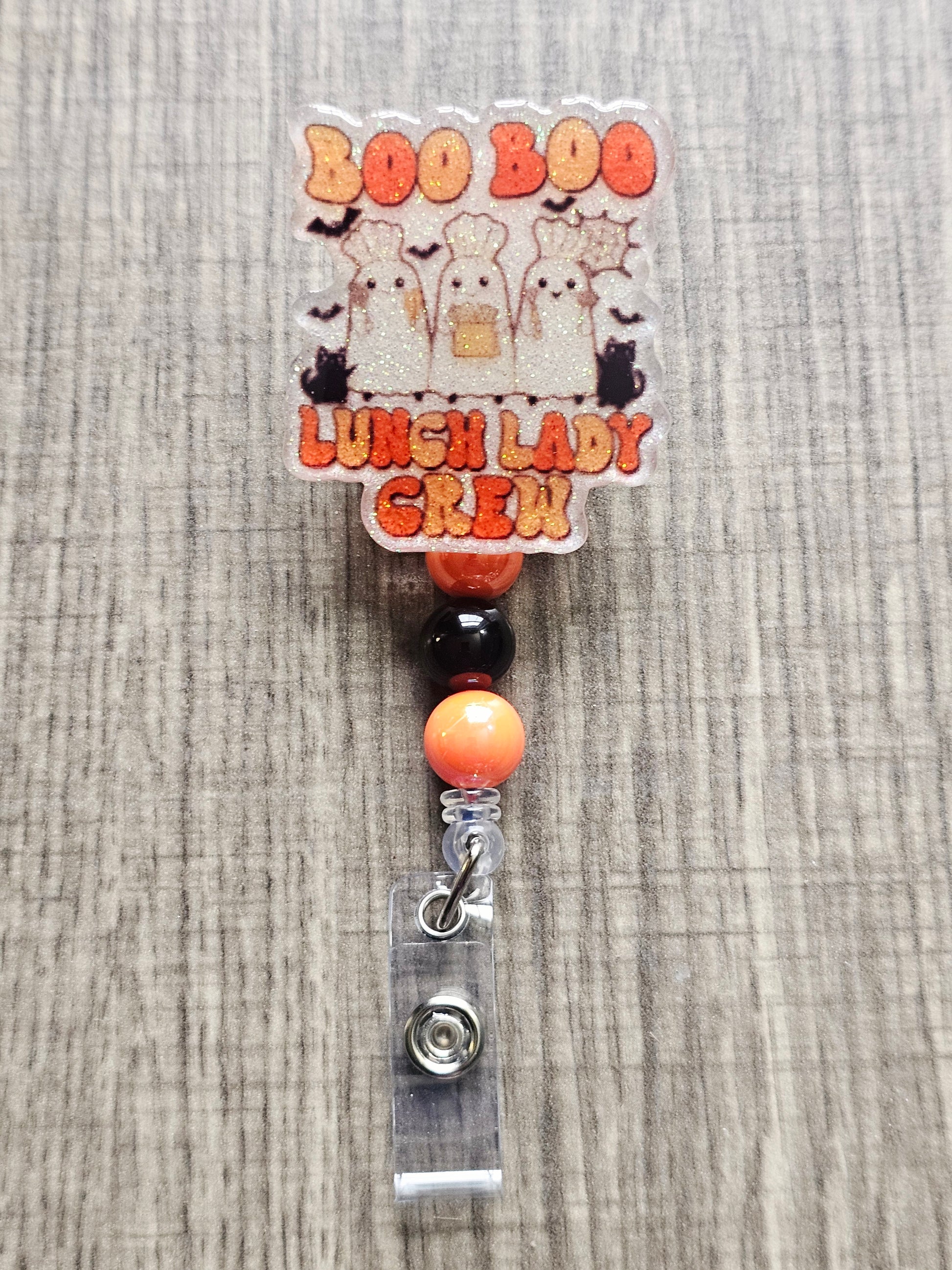 White glitter acrylic badge reel with "Boo Boo Lunch Lady Crew" in a retro Halloween theme, featuring three ghost lunch ladies in chef hats with black cats, bats, and cooking utensils, with matching decorative beads.