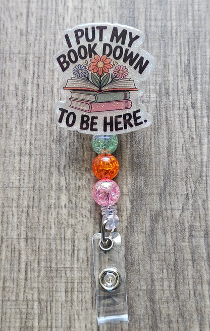 Retractable book-themed badge reel featuring a stack of colorful books, an open book with blooming flowers, and text that reads "I Put my book down to be here." 