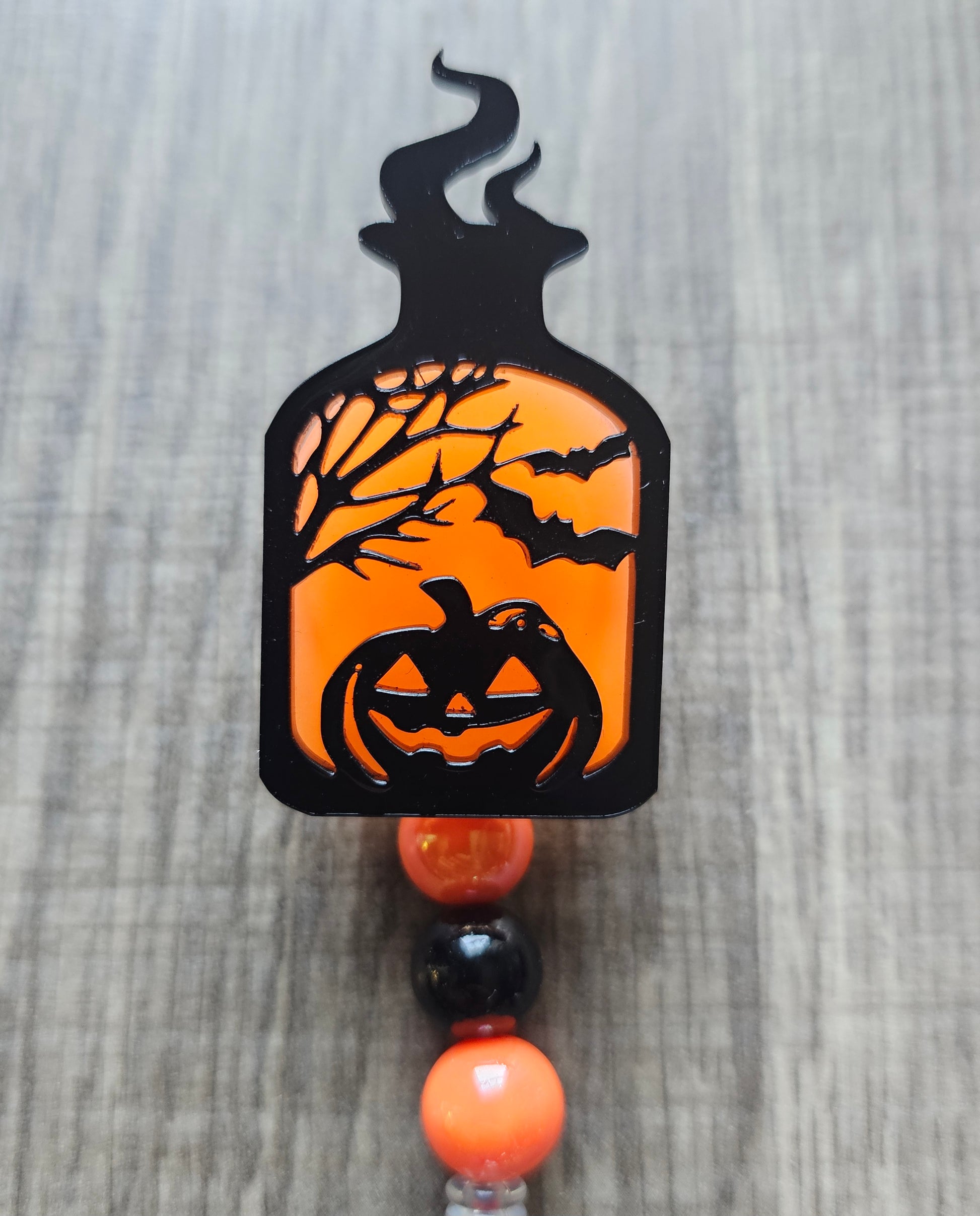 Orange glow-in-the-dark potion bottle-shaped badge reel with an eerily smiling jack-o-lantern surrounded by trees and bats, with matching glow-in-the-dark decorative beads.