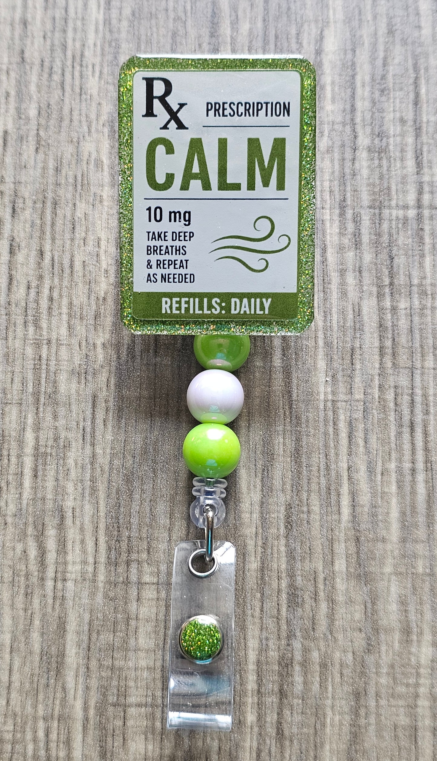 A green and white 'Calm' 10 mg prescription bell bottle with a beaded strap and clear acrylic tag, displayed on a gray wood-grain background.