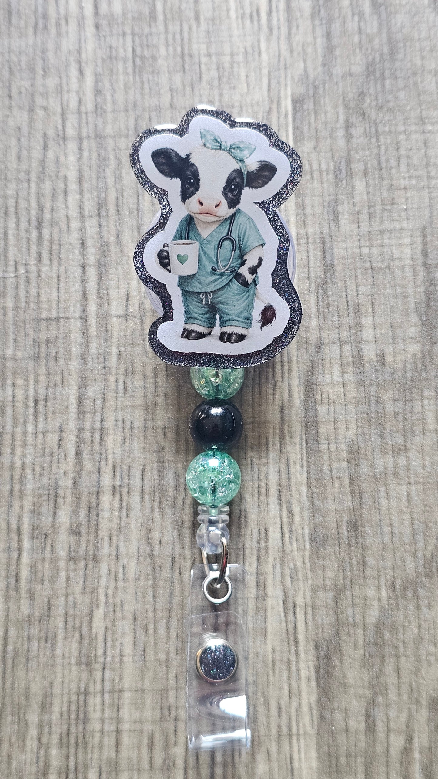 A custom beaded retractable badge reel featuring a die-cut sticker of a cartoon cow dressed as a doctor in teal scrubs, displayed on a light wood grain background.
