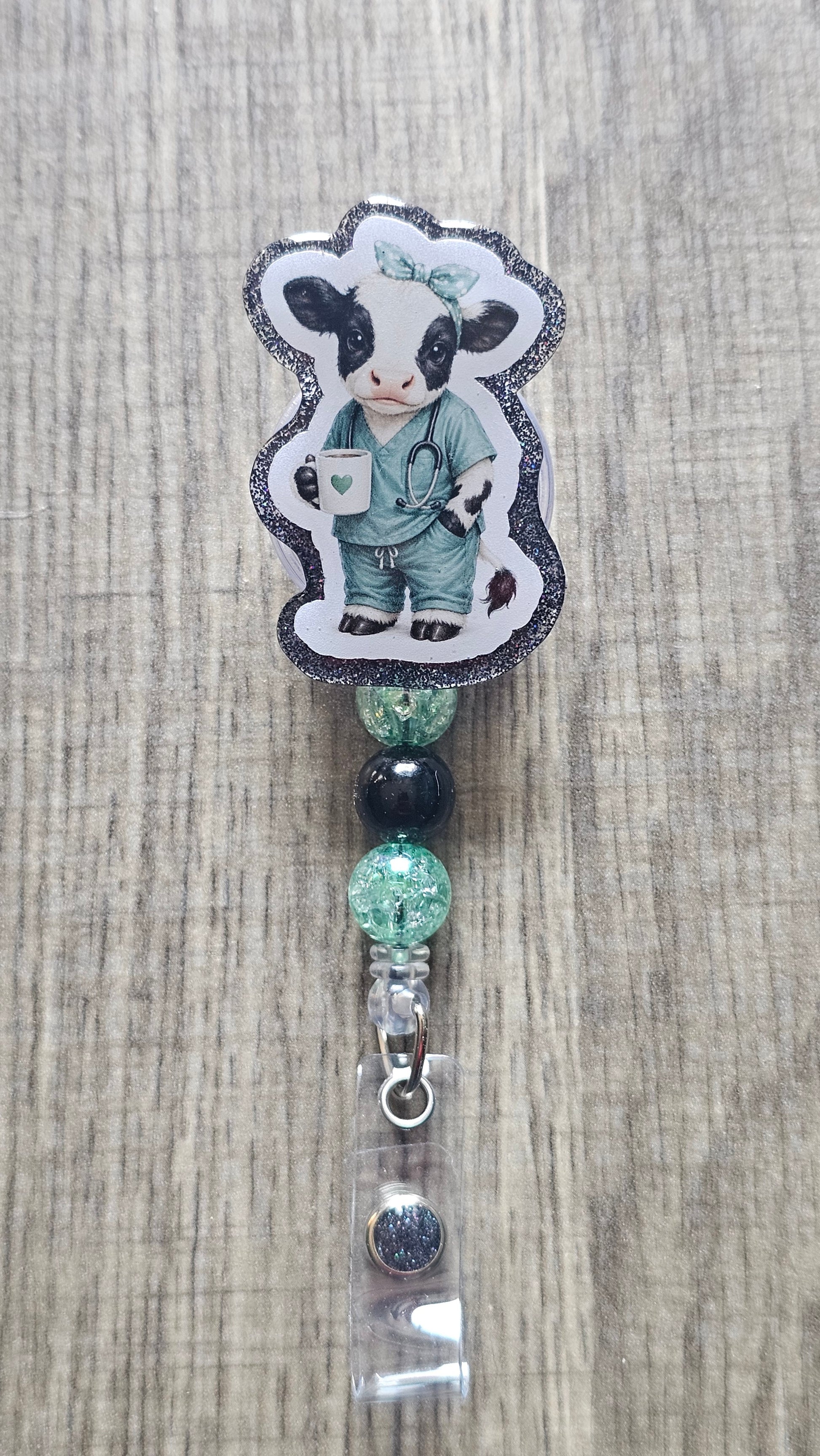 A custom beaded retractable badge reel featuring a die-cut sticker of a cartoon cow dressed as a doctor in teal scrubs, displayed on a light wood grain background.