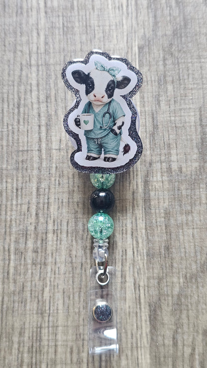 A custom beaded retractable badge reel featuring a die-cut sticker of a cartoon cow dressed as a doctor in teal scrubs, displayed on a light wood grain background.
