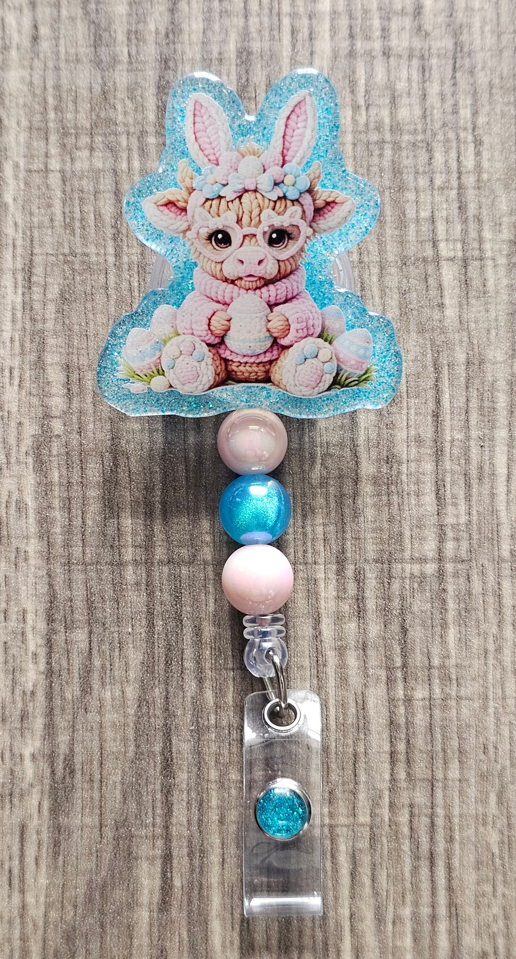 Close-up of a crocheted highland cow badge topper wearing pink bunny ears and holding a blue Easter egg.