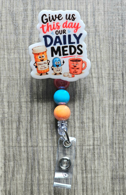 Cartoon coffee mug, pill bottle, and capsule with the text "Give us this day our daily meds" for a retractable badge reel.