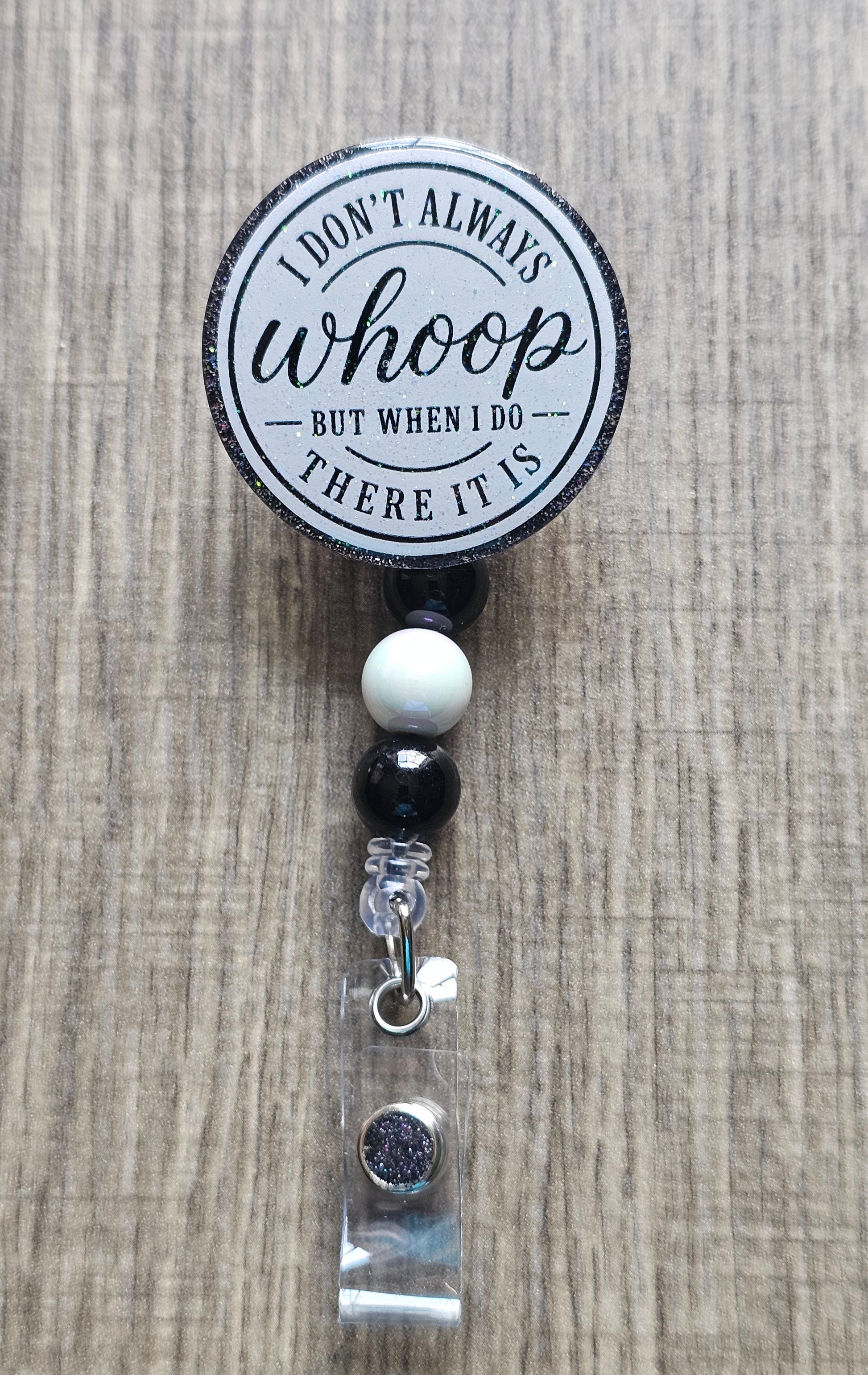 A retractable badge reel with a circular black and white printed top that reads 'I DON'T ALWAYS whoop BUT WHEN I DO THERE IT IS', shown on a light wood background. The reel is attached to black and white beads, a clear acrylic sleeve, and a silver lobster clasp.
