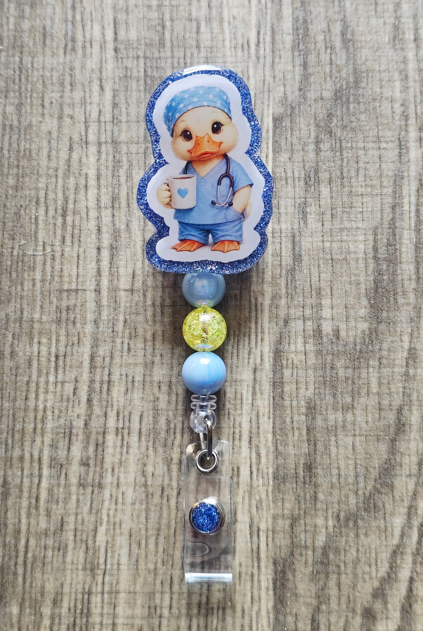 A baby nurse-themed bouncer clip or roll bar accessory featuring a cartoon duck character in blue scrubs, blue glitter trim, and decorative beads on a wood background.