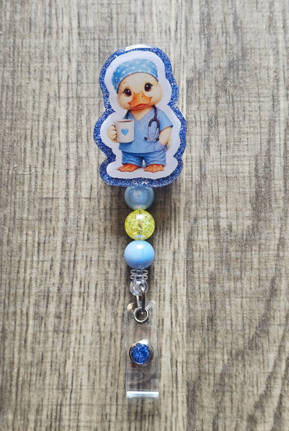 A baby nurse-themed bouncer clip or roll bar accessory featuring a cartoon duck character in blue scrubs, blue glitter trim, and decorative beads on a wood background.