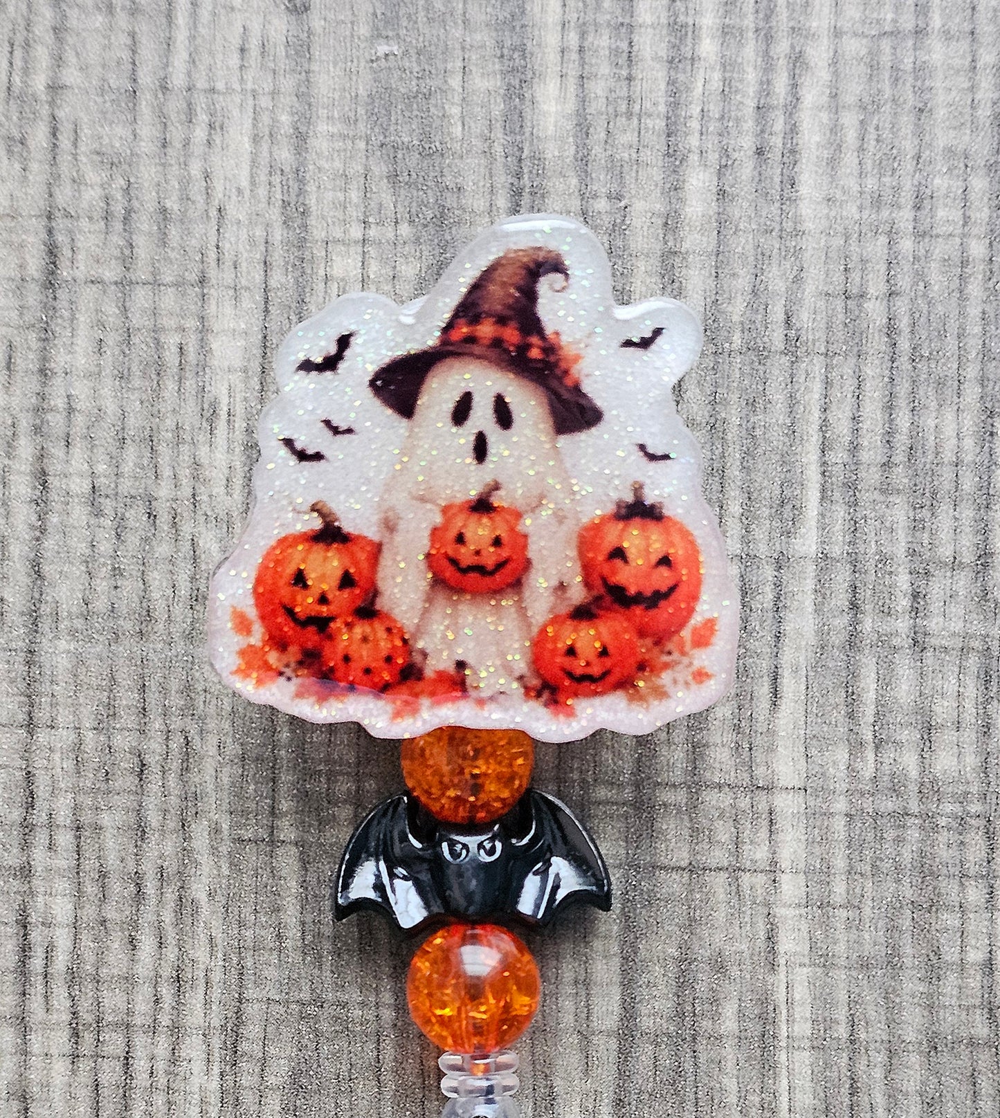 Halloween badge reel with a ghost holding a jack-o-lantern bucket in a pumpkin patch, styled with a bat-shaped decorative bead.