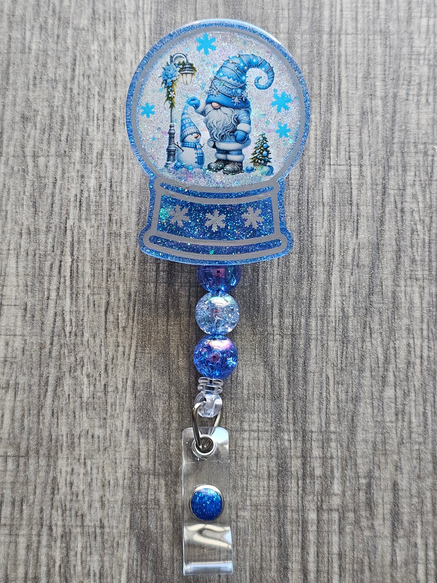 A badge reel with a snow globe design featuring a winter Gnome surrounded by snow and a decorated lamp along a snowman. 