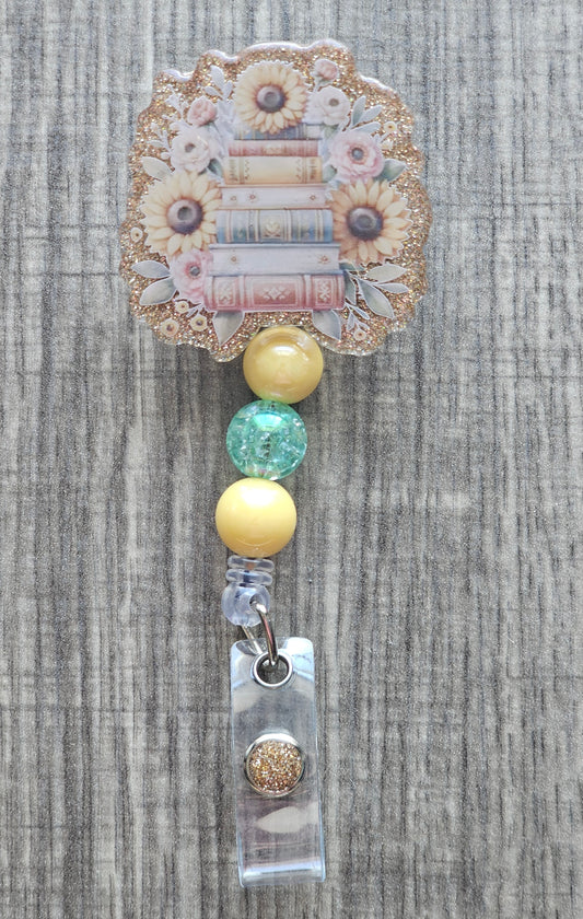 Retractable badge reel showing a stack of books with sunflowers on a gold background with matching yellow beads. 