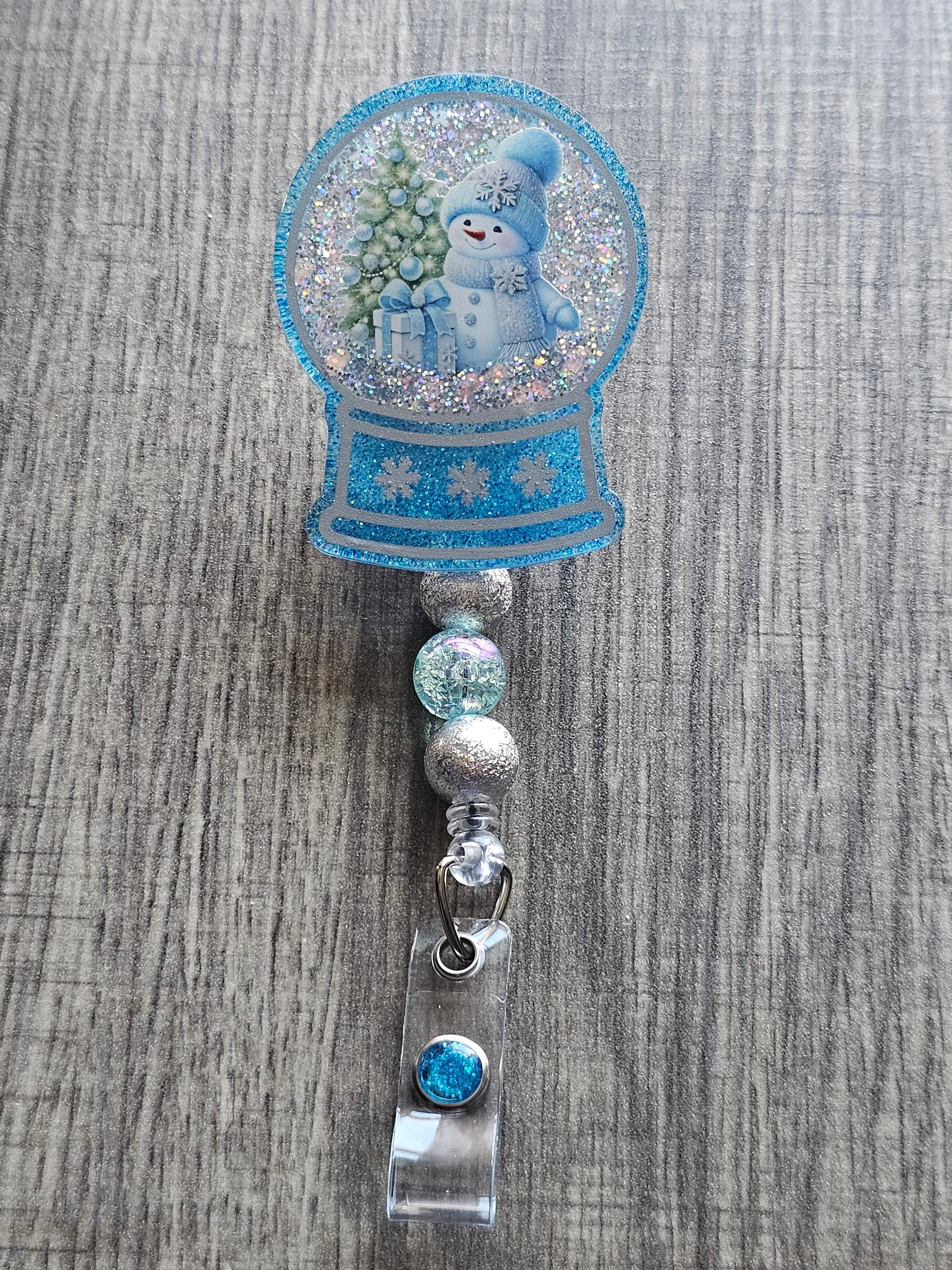 A badge reel with a snow globe design featuring a snowman in a hat  and scarf with a Christmas tree near him along with a Christmas present in front of him. 