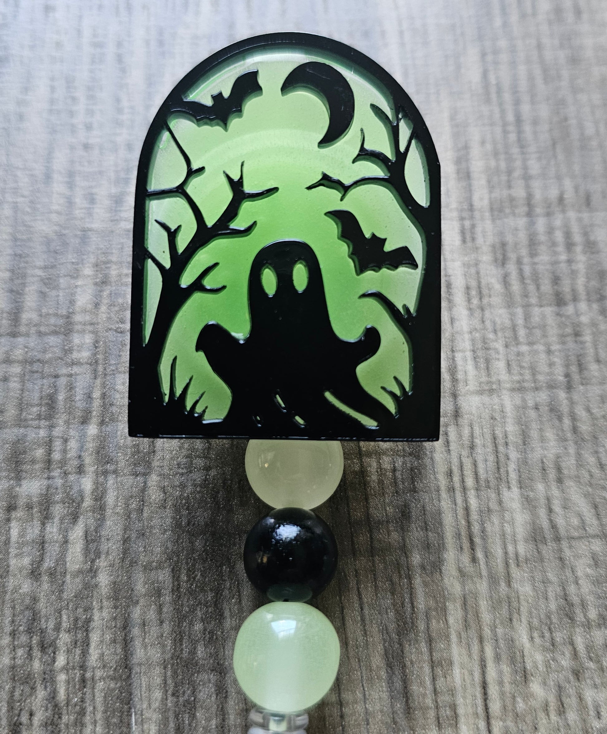 Green glow-in-the-dark tombstone-shaped badge reel with an eerie ghost emerging from the ground, surrounded by trees, a moon, and bats, with matching glow-in-the-dark decorative beads.