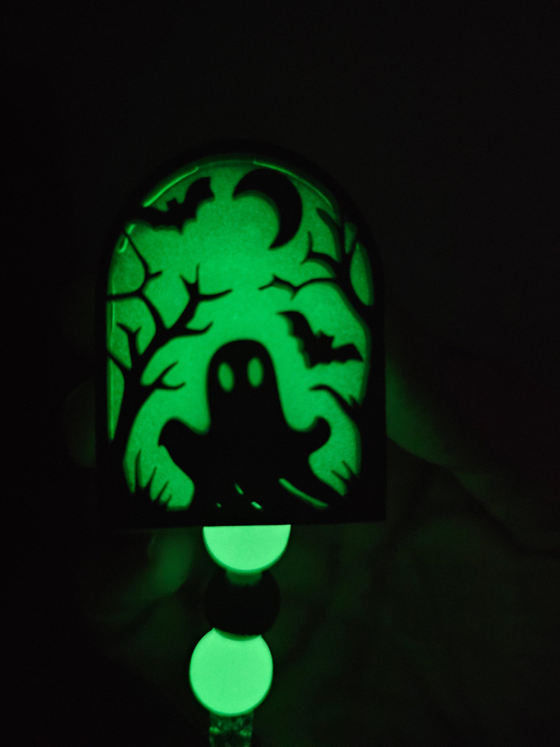 Green glow-in-the-dark tombstone-shaped badge reel with an eerie ghost emerging from the ground, surrounded by trees, a moon, and bats, with matching glow-in-the-dark decorative beads.