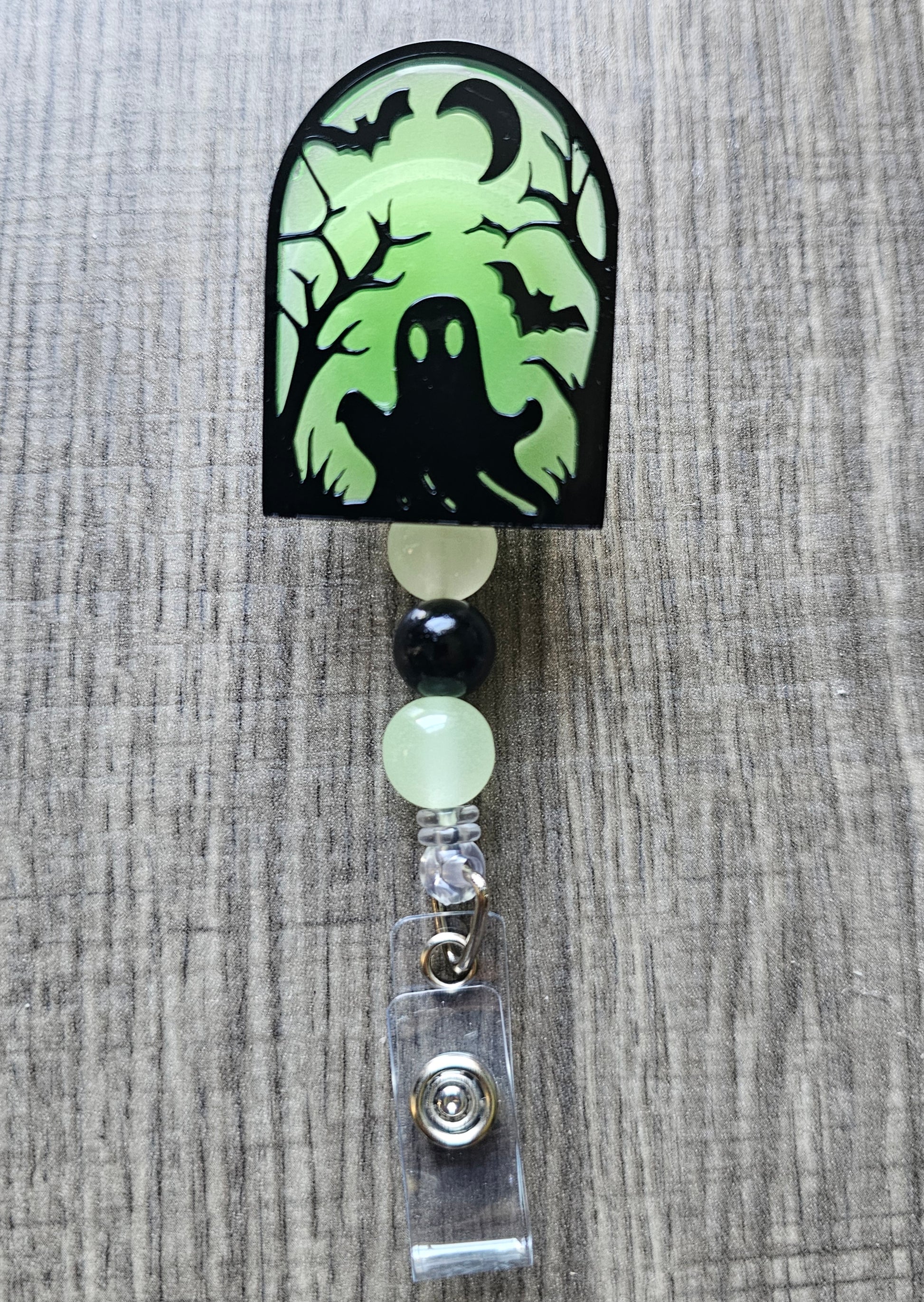 Green glow-in-the-dark tombstone-shaped badge reel with an eerie ghost emerging from the ground, surrounded by trees, a moon, and bats, with matching glow-in-the-dark decorative beads.