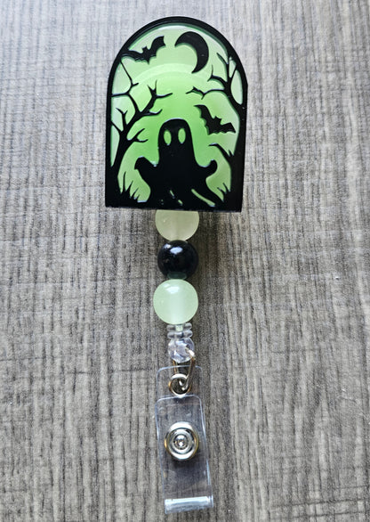 Green glow-in-the-dark tombstone-shaped badge reel with an eerie ghost emerging from the ground, surrounded by trees, a moon, and bats, with matching glow-in-the-dark decorative beads.
